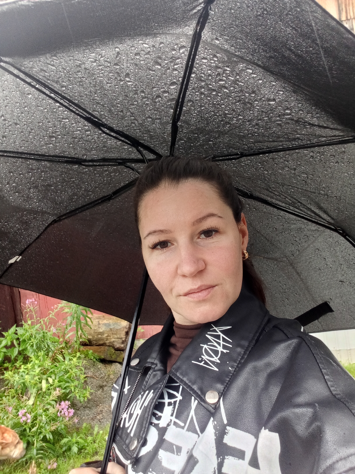A young woman with dark hair pulled back stands outdoors under a black umbrella, which is covered in raindrops. She is wearing a dark brown turtleneck shirt and a black faux leather jacket with white graphic designs and text. She has on a gold hoop earring in her visible ear. The background shows greenery, a red wooden structure, and some rocks. The scene appears to be in a rural or suburban area, potentially in Zlatoust, Russia, given the context provided.