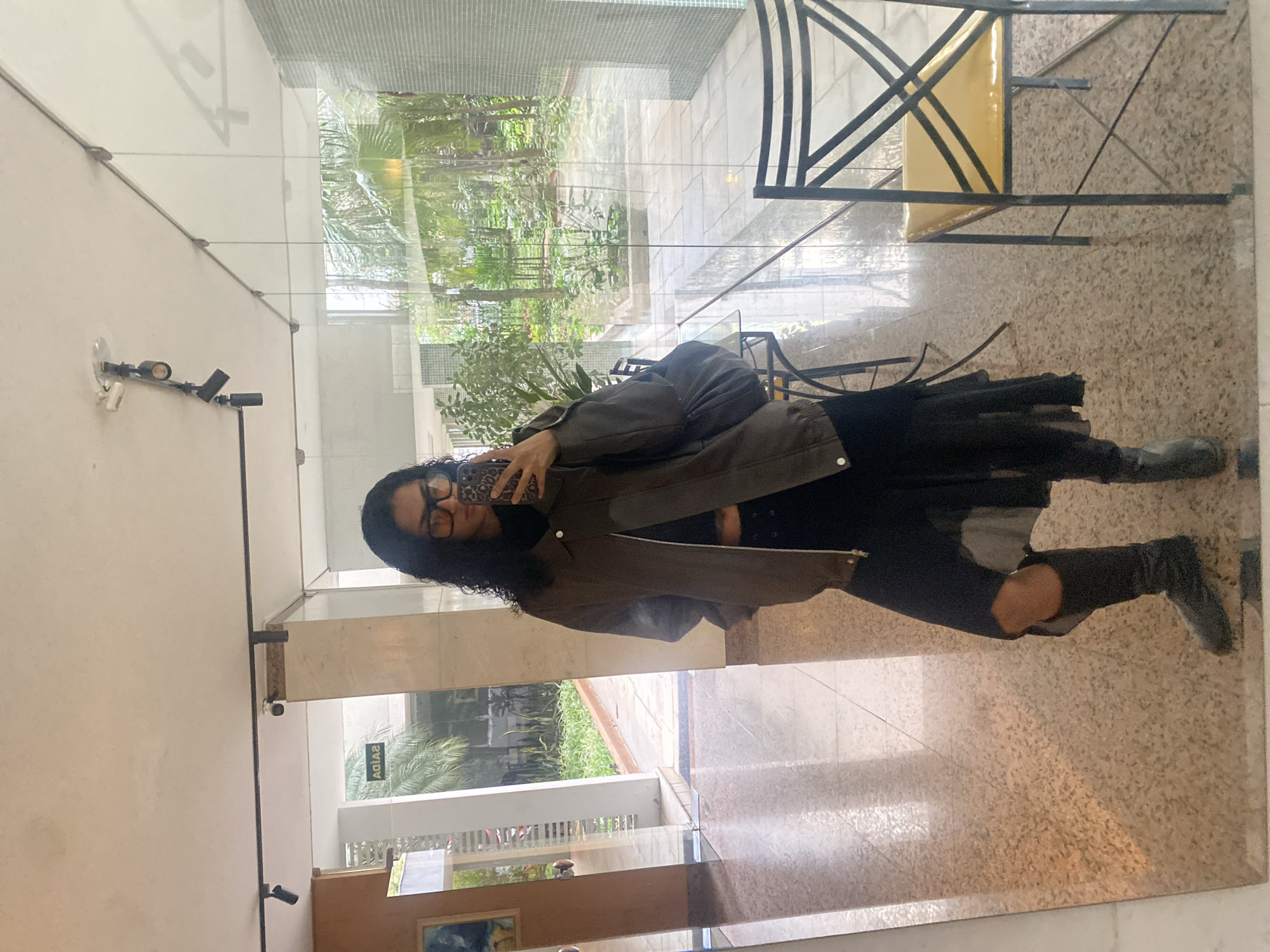A person with dark curly hair and glasses is taking a selfie in front of a mirrored wall. The person is wearing a brown jacket, black top, and dark jeans with a hole in the knee. They are holding a phone with a patterned case.
The reflection shows a bright environment with a green outdoor space visible through a glass wall. A metal chair is also reflected in the mirror.
The location is likely in São Paulo, Brazil, indicated by the word "SAÍDA" (Exit in Portuguese) on a mirrored sign on the wall in the reflection. The indoor setting appears to be a lobby or entrance area.