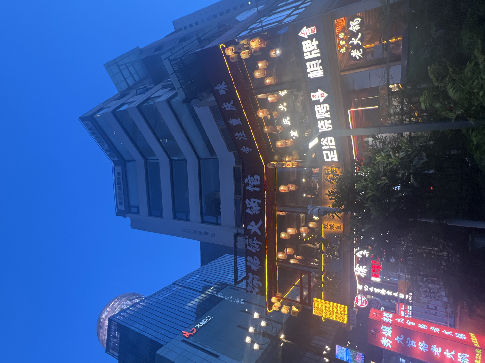 The image depicts a nighttime view of a city street in Yuzhong District, China, with a clear blue sky overhead. Dominating the scene are several buildings of varying heights and architectural styles. On the left, a modern skyscraper with a reflective glass facade rises, topped by a large, spherical, illuminated structure. Adjacent to it, a darker, taller building features rows of illuminated windows. To the right, a multi-story building with a stepped facade is visible.
Below these structures, a vibrant commercial area is active. A prominent red sign with Chinese characters, likely advertising a restaurant, is lit up, accompanied by numerous red lanterns strung across the facade. Below this, another row of illuminated signs in Chinese characters advertise various services, including "Foot Bath," "Barbecue," and "Hot Pot." Some of these signs are vertical and neon.
In the foreground, lush green foliage partially obscures the lower parts of the buildings and signs. The street itself is not directly visible, but the illuminated signs suggest a bustling urban environment. The overall impression is one of a lively commercial district at night.
