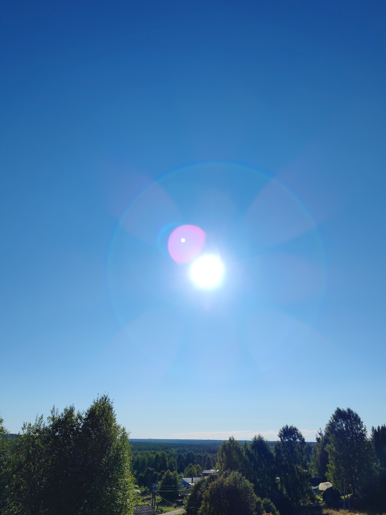 The image depicts a clear blue sky with the sun prominently visible, exhibiting lens flare. Below the sky, a landscape stretches out, characterized by a dense forest that extends to the horizon. In the foreground, several trees are visible, with some appearing larger and closer to the viewer. The lower portion of the image shows glimpses of a rural settlement, with scattered buildings and what appears to be a road or path. The overall scene suggests an outdoor, natural environment with signs of human habitation. The location is specified as Чухлэм, Russia.