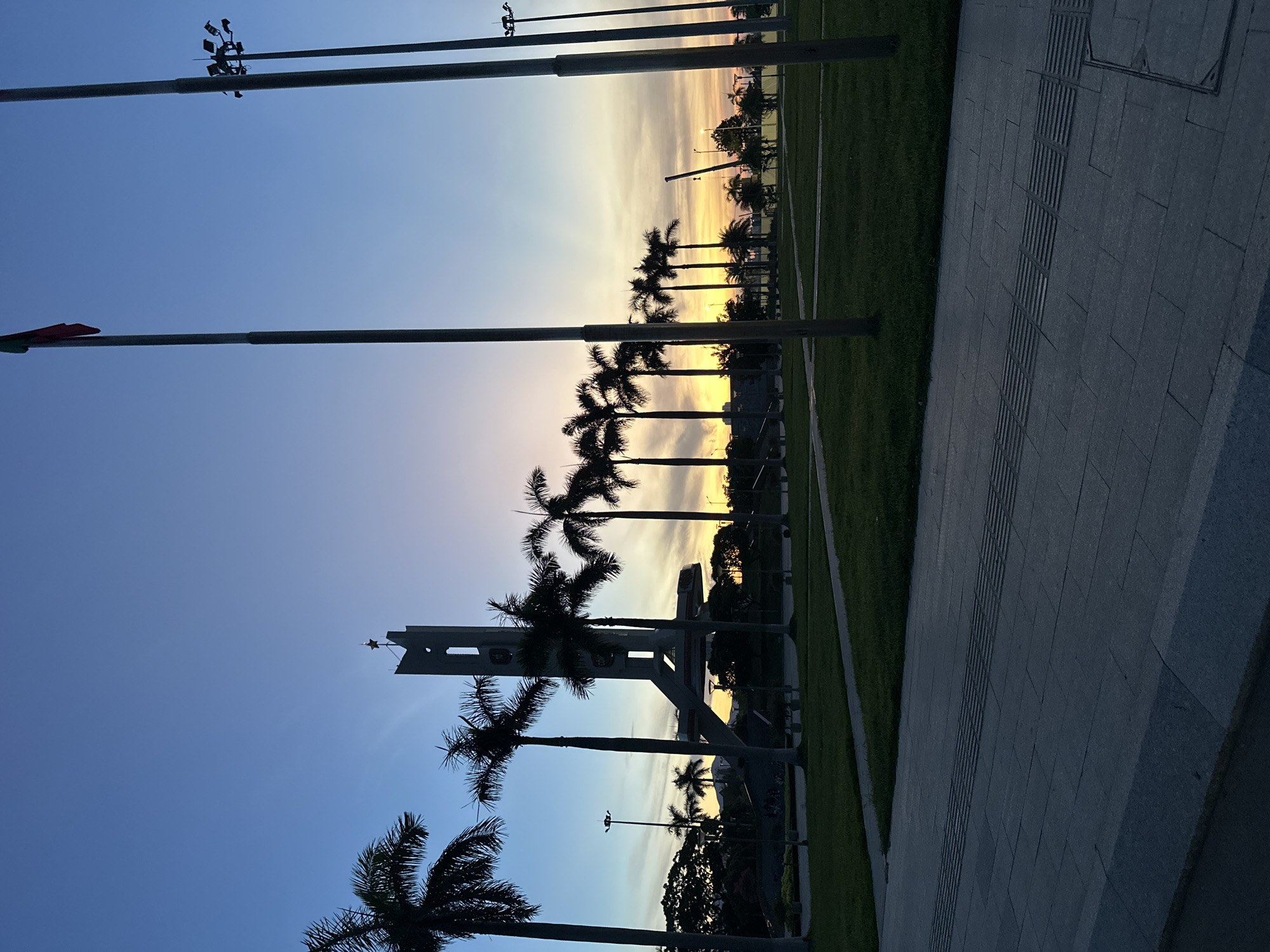 The image depicts a sunset scene at the Dragon Bridge in Da Nang, Vietnam. The Dragon Bridge is prominently featured in the background, with its iconic structure silhouetted against the colorful sky. Several palm trees line the foreground and middle ground, their fronds casting shadows on the manicured lawn. A paved walkway with tactile paving runs along the bottom of the image. The sky displays a gradient of blue and orange hues, indicative of the setting sun. There are no people visible in the image. The overall scene is serene and picturesque, capturing the architectural beauty of the Dragon Bridge at dusk.