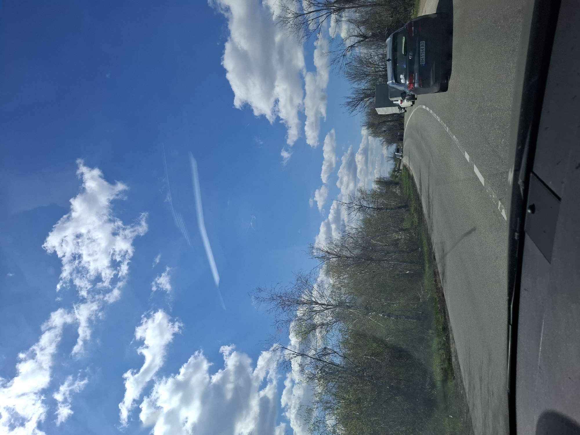 The image depicts a road scene likely taken from inside a moving vehicle in Tsar Simeonovo, Bulgaria. A dark grey Hyundai car with license plate "BH1164 AO" is visible ahead on the road, followed by what appears to be a small trailer. The road curves gently, with trees and vegetation lining its side. The sky is blue with scattered white clouds. The image is somewhat distorted, likely due to the angle of the camera.