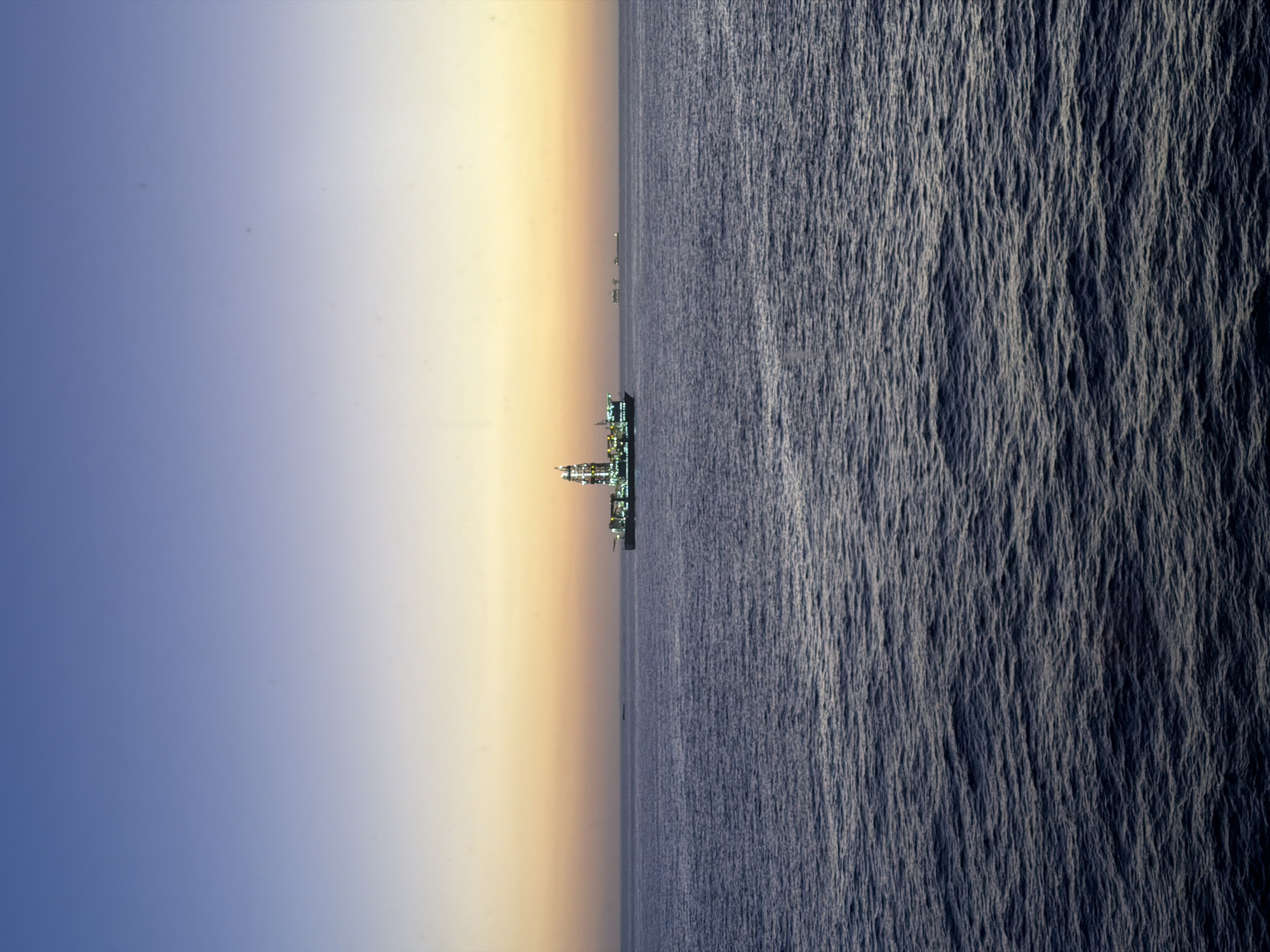 The image captures a vast seascape at either dawn or dusk, with a clear horizon dividing the sky from the ocean. The sky, occupying the left half of the frame, displays a striking gradient from a deep, clear blue at its upper extent to a warm, soft yellow and orange closer to the horizon.
Centered on the horizon line is a large, brightly illuminated offshore platform or industrial vessel, possibly an oil rig or a large ship, with numerous white and green lights clearly visible against the low light of the sky. Several smaller, less distinct structures or vessels are also visible further along the horizon in the distance.
The right half of the image is dominated by the deep blue ocean, characterized by a textured surface of countless small ripples and waves, suggesting calm to moderately choppy waters. There are no people or visible text in the scene. The overall impression is one of an isolated industrial presence against the backdrop of a grand natural environment.
