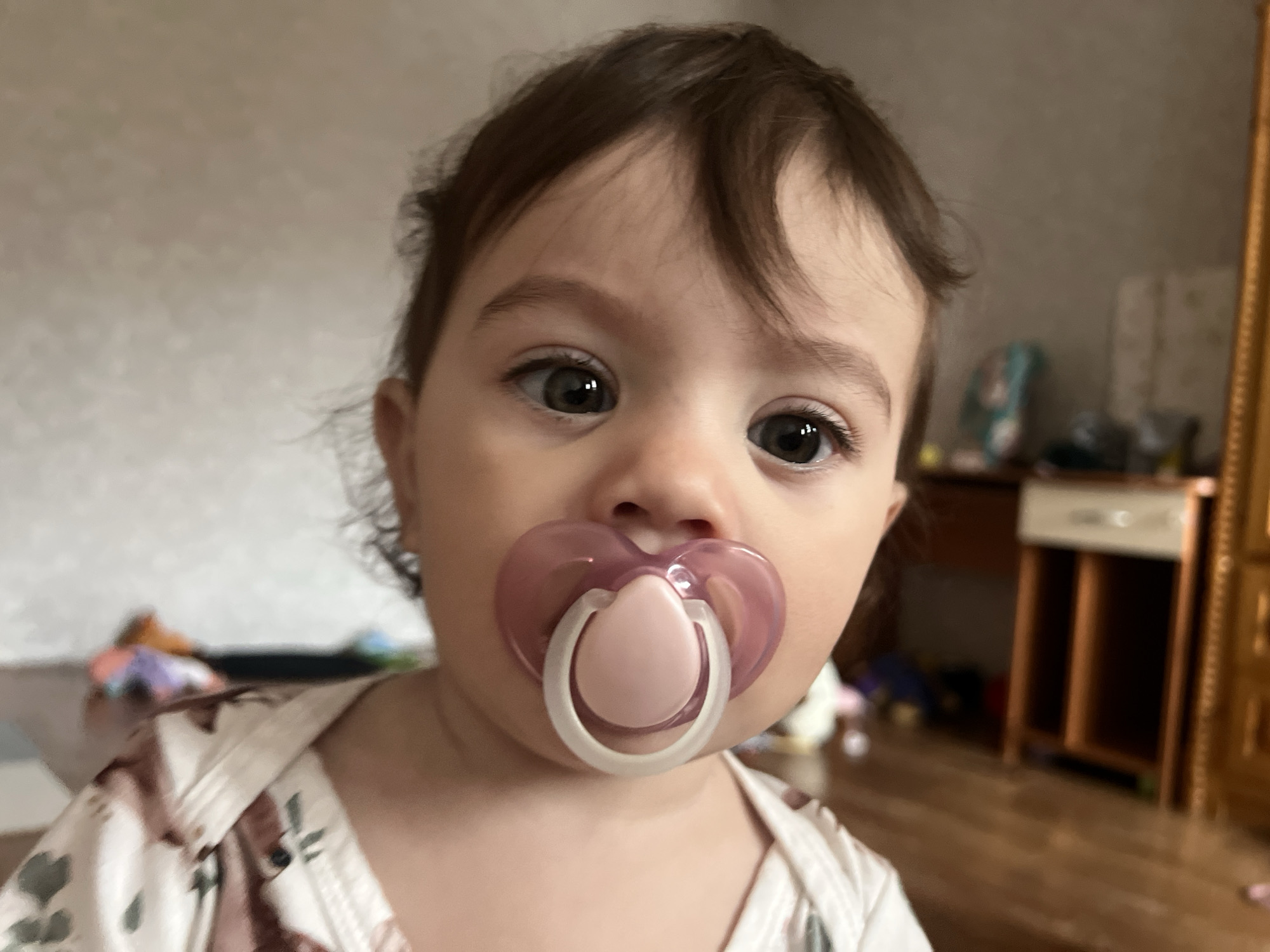 A close-up, eye-level shot of a baby with a pacifier in its mouth. The baby has brown hair and eyes, and light skin. The pacifier is pink and white. The baby is wearing a white shirt with a floral pattern. The background is blurry and shows a room with a desk and a wooden cabinet. The image was likely taken indoors in Elena, Bulgaria.