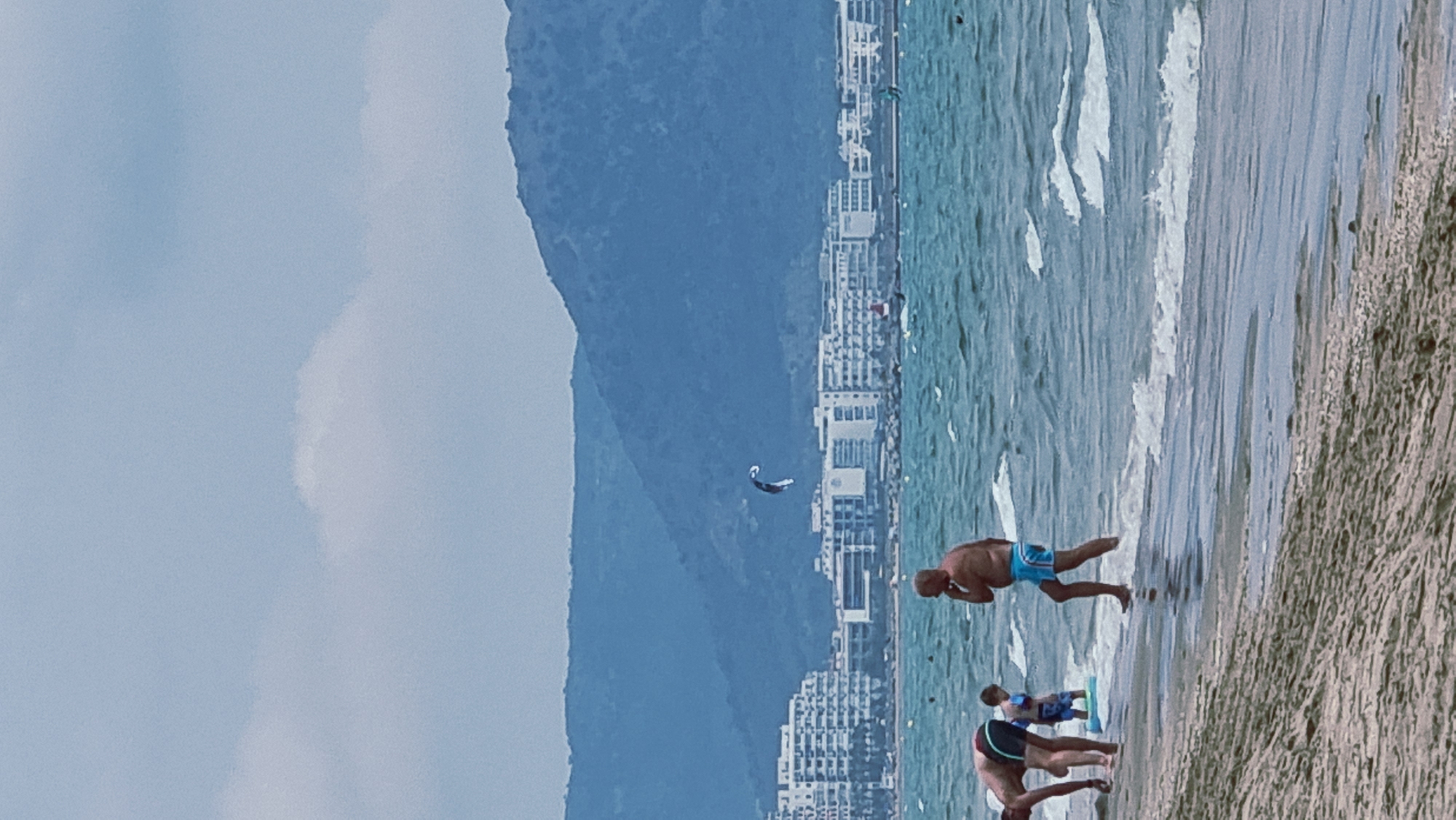 The image captures a scene at Empuriabrava beach in Castelló d'Empúries, Spain. The sky is overcast with scattered clouds. In the background, a line of multi-story white buildings forms the coastline. Beyond the buildings, a large, dark, mountainous terrain is visible. The sea is a vibrant turquoise with moderate waves. A kite surfer is in the air over the water.
In the foreground, on the sandy beach, several individuals are present. A man in blue swim trunks is standing in the shallow water, looking to his left. To his left, a younger person in blue swim trunks stands on a light blue float. Further to the left, a person with shorts is bent over. The beach itself is composed of sand with some debris. The overall impression is a typical summer day at a coastal resort.