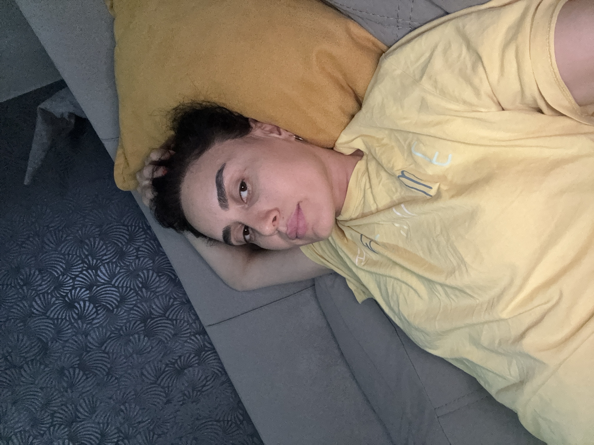 A woman is lying on a couch, resting her head on a yellow pillow. She is wearing a yellow t-shirt and her left arm is behind her head. The background shows a dark grey textured wallpaper and a grey couch. The image is a selfie taken from a high angle, showing the woman's face and upper body. The setting is indoors, likely a living room. The image does not provide any information about the city of Tbilisi, Georgia.