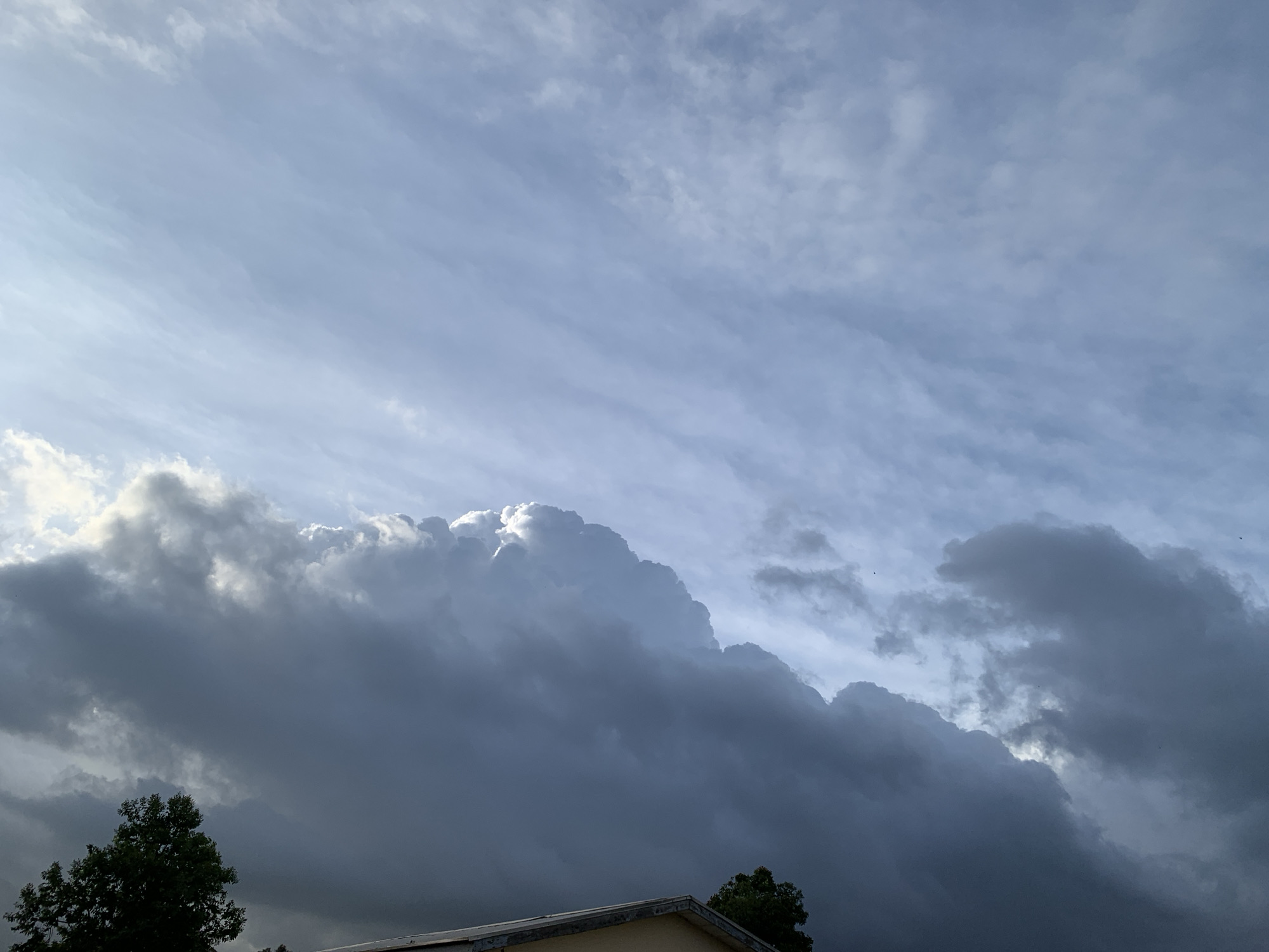 The image depicts a sky filled with a dramatic interplay of light and shadow created by clouds. In the foreground, the dark, heavy clouds appear to be moving in, with a few trees and the edge of a building visible at the bottom. The clouds are mostly dark gray and blue, but a bright, illuminated cloud formation is prominent in the center of the image, suggesting sunlight breaking through. The sky above is a lighter blue with wispy, streaky clouds that are more translucent. The overall impression is of an impending storm or the aftermath of one, with a sense of dynamic weather. There is no visible indication of the city of Kuru Karama, Nigeria. The scene focuses solely on the sky and the very tops of some natural and man-made structures.