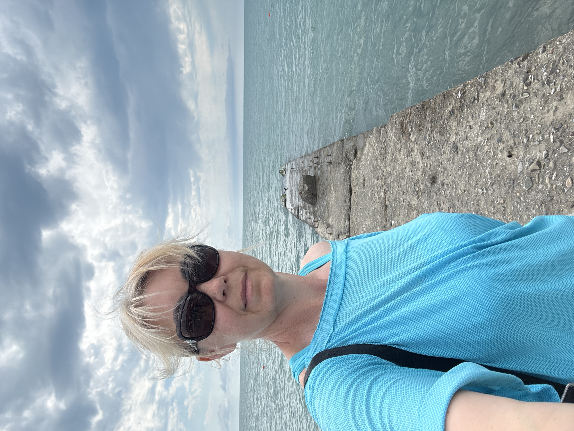 The media file is a portrait-oriented selfie taken by a woman standing on a concrete pier by the water. She has blonde hair and is wearing sunglasses and a bright blue athletic top. The sky is cloudy, suggesting it might be an overcast day. The water appears calm and a light blue-green color, with a single red marker visible in the distance. The concrete pier is rough and weathered, with some visible rebar and pebbles. The background shows the vast expanse of the water meeting the horizon. The woman's expression is neutral. The image appears to have been taken during daylight hours. There is no visible text in the image. Given the location context of Sochi, Russia, this could be a view of the Black Sea.