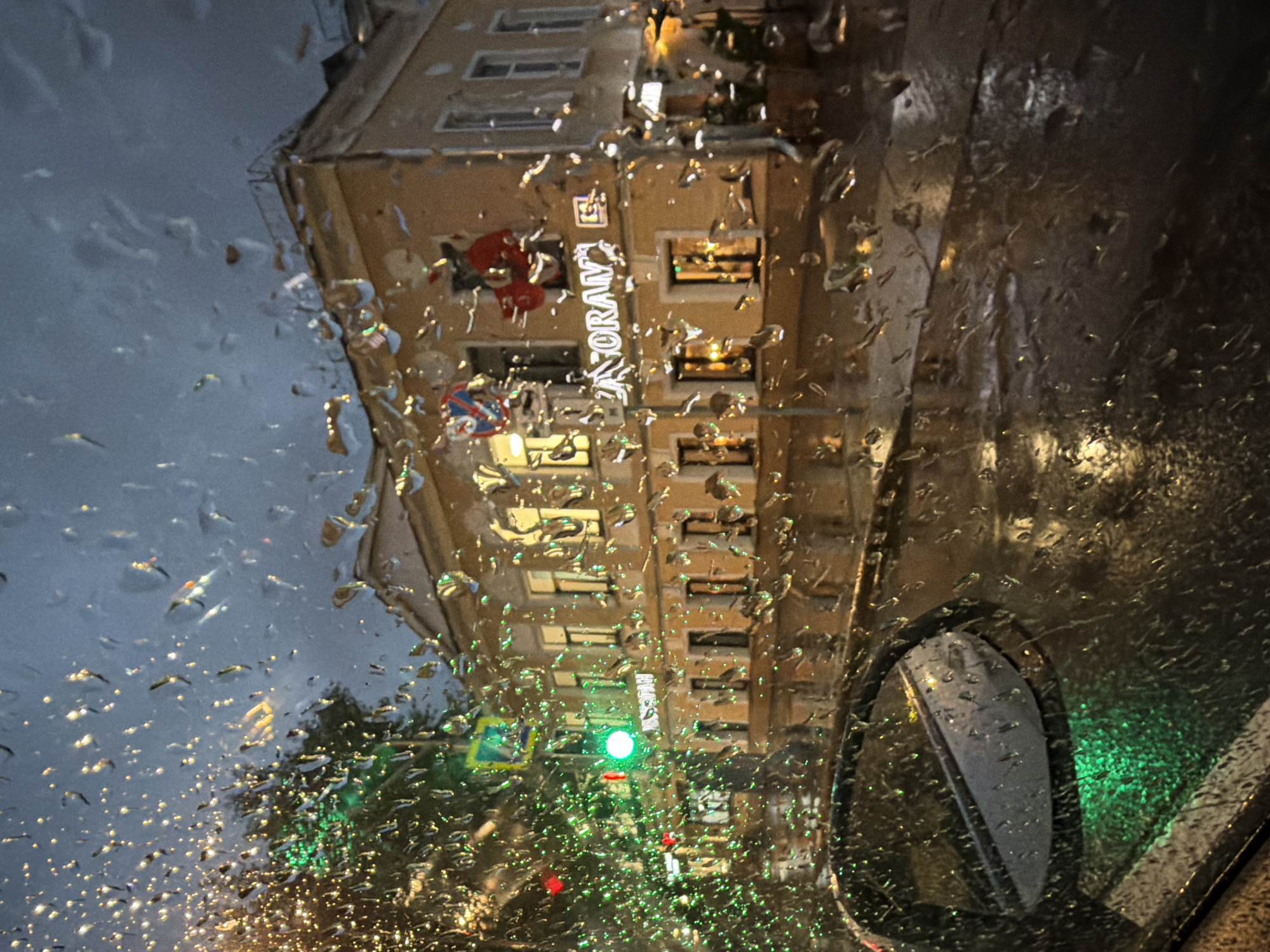 The image depicts a street scene at night in Moscow, Russia, viewed through a rain-streaked window, likely from inside a vehicle. The foreground is dominated by numerous raindrops on the glass, distorting the view of the city beyond. To the left, a car's side mirror is visible, reflecting a green light. A traffic light displaying green is also present, along with red lights further in the distance. A blurry green Christmas tree is visible on the left side of the frame.
In the mid-ground, a multi-story building with illuminated windows stands. The building is light yellow and has several floors. A sign on the building reads "RESTORAN" in white illuminated letters. A red decorative element, possibly a large ornament or a stylized figure, is mounted above the sign. A circular sign with a red border and a white design is also visible on the building.
The street and sidewalk are wet and reflective due to the rain, with lights from the building and streetlights creating reflections on the surface. The sky is dark and overcast, indicative of nighttime and rainy weather. No people are clearly discernible in the image. The overall impression is a melancholic and atmospheric view of a city in the rain.