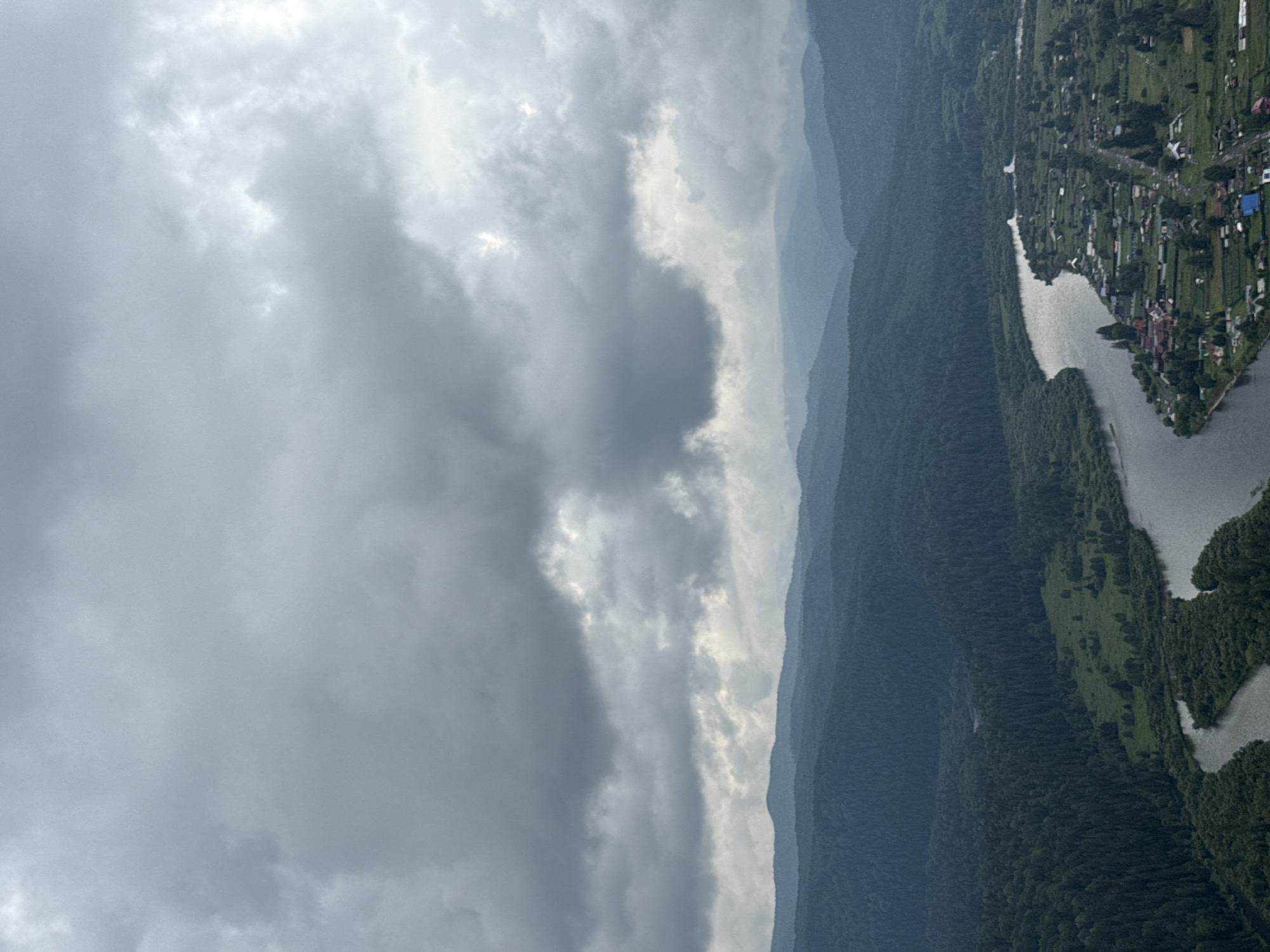 An aerial, wide-angle, daylight photograph captures a panoramic view of a rural landscape in Urman, Russia. The sky is overcast with heavy, dark grey clouds that dominate the upper half of the frame. Below the clouds, layers of dark green, tree-covered mountains recede into the distance, their peaks softened by atmospheric haze. In the foreground, a wide, grey river snakes through a lush green valley. On the right bank of the river, a village is nestled amongst the trees, with numerous small houses, some with red roofs, scattered across the landscape. A road runs through the village, and smaller waterways branch off from the main river. The overall scene depicts a remote, natural environment with a small human settlement integrated into it.