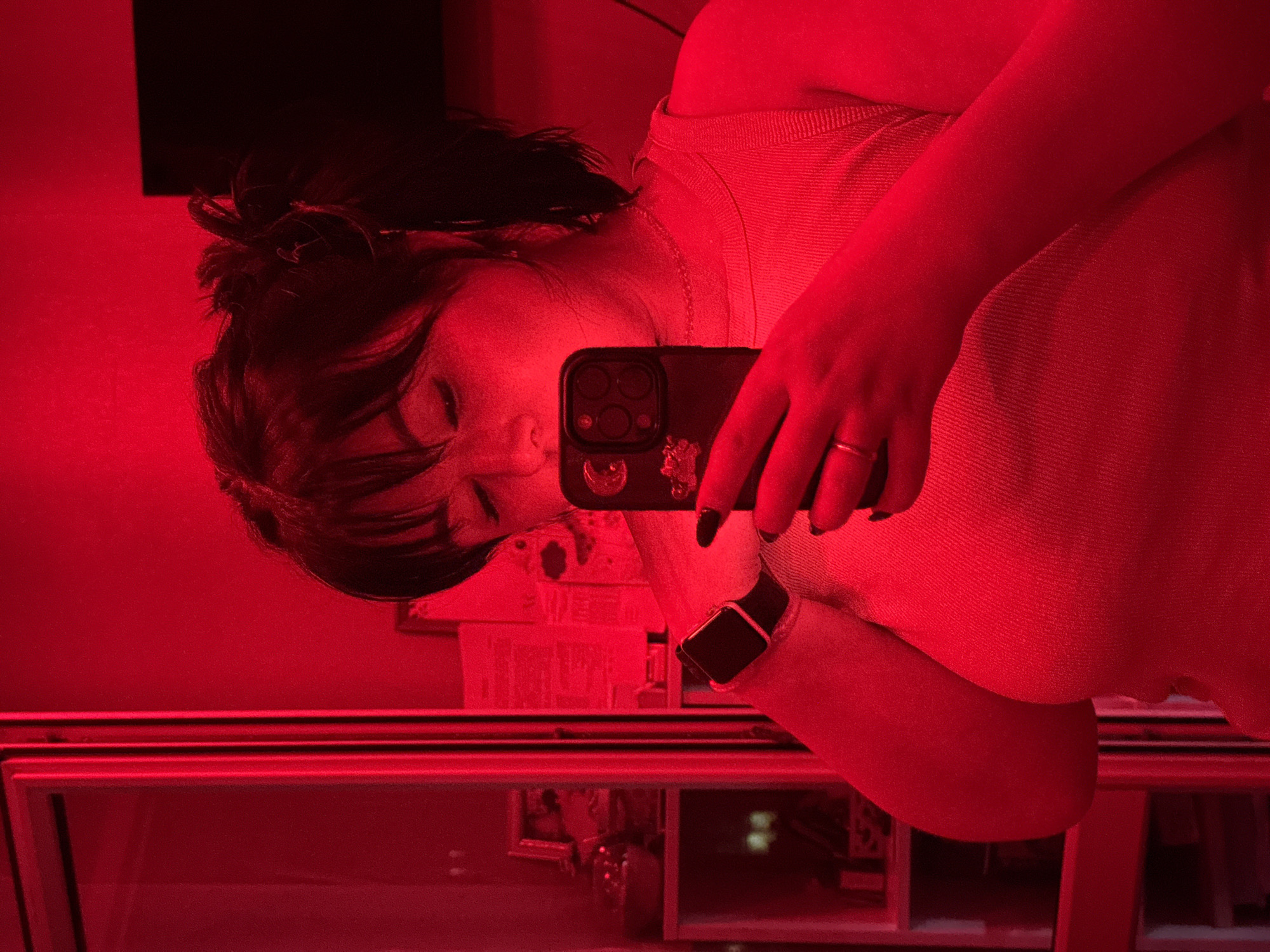 A person with dark hair pulled back and bangs is holding a phone to take a selfie in a mirror. The phone case is dark colored with embellishments on it. The person is wearing a light-colored top and a smartwatch on their left wrist. The reflection in the mirror shows a shelving unit or display case. The lighting in the room is predominantly red. While the exact location in Chaikovsky, Russia is not discernible from the image, the presence of a display case suggests the person is in a commercial establishment, possibly a store or restaurant.