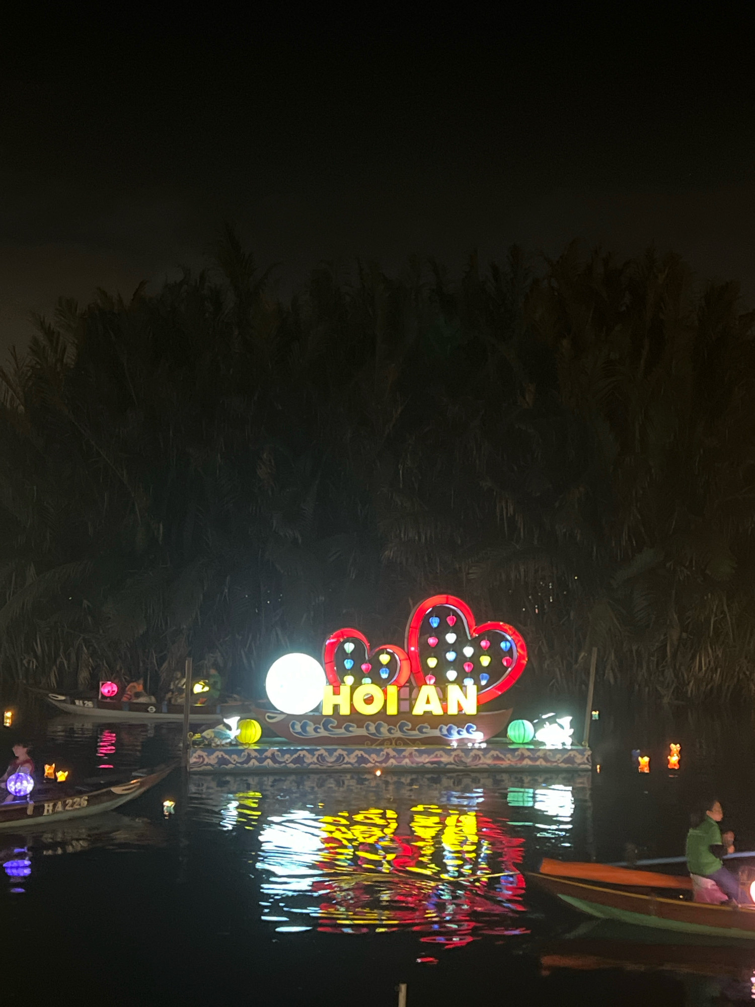 At night in Hoi An, Vietnam, a boat floats on a dark river, illuminated by a vibrant display of colorful lanterns. The central focus is a boat adorned with a large, illuminated sign spelling "HOI AN" in bright yellow letters. This sign is framed by two prominent heart shapes, outlined in red and filled with small, colorful heart-shaped lanterns. To the left of the "HOI AN" sign, a large, bright white sphere lantern adds to the illumination. The boat itself features decorative blue and white wave patterns along its edge.
Several other boats are visible on the river. One boat in the foreground to the left carries a person holding a purple spherical lantern. Another boat further back also has people and is decorated with pink and yellow lanterns. To the right, a boat contains a person wearing a green shirt, holding a lit lantern. Smaller, individual lanterns, some appearing to be in the shape of teddy bears, float on the water's surface and are attached to other boats, casting reflections. The background is dominated by dark foliage, likely palm trees, which are silhouetted against the night sky. The water's surface reflects the bright lights of the lanterns, creating a dazzling display of colors.