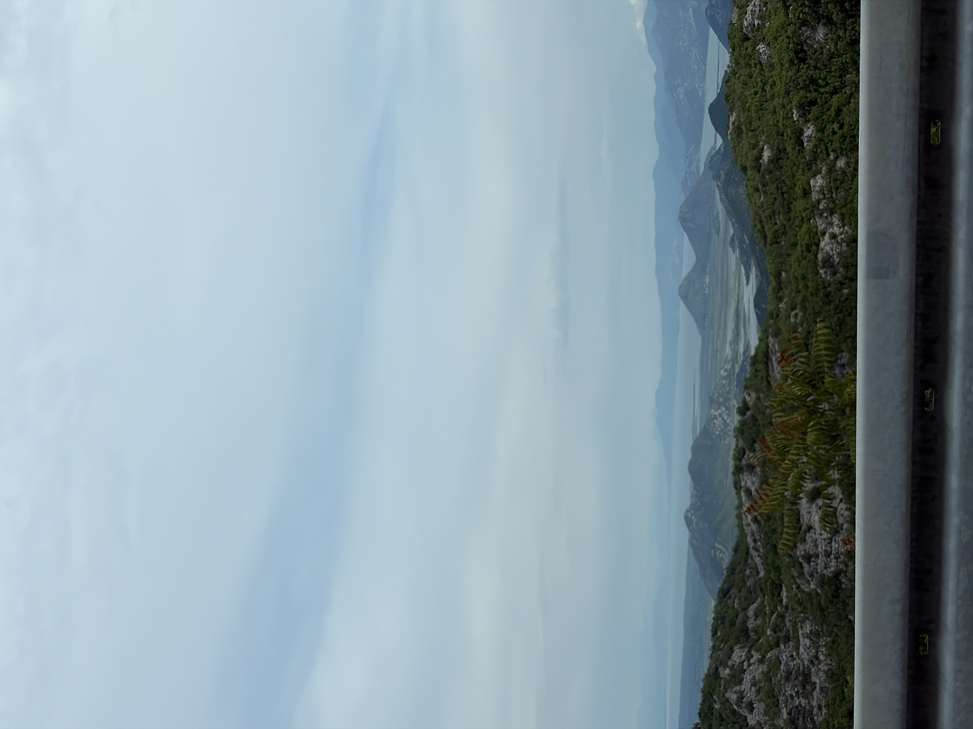 The image captures a vista from what appears to be a roadside in the vicinity of Gornja Brova, Montenegro. To the right, a gray metal guardrail runs vertically, its texture and details visible. Beyond the rail, the landscape slopes down, covered in lush green vegetation with rocky outcrops. In the distance, a series of mountains stretch out, shrouded in a light haze. Water bodies snake between the mountains, reflecting the sky. The sky itself is overcast, with soft blue hues peeking through the clouds.