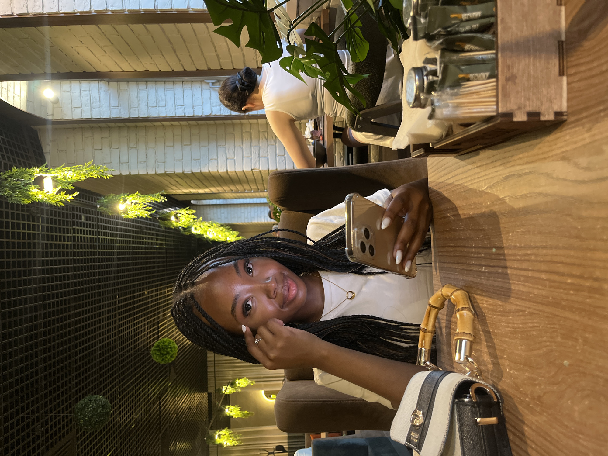 The image depicts a young Black woman with braided hair sitting at a wooden table in what appears to be a cafe or restaurant. She is looking at the camera and holding a smartphone. To her left, a purse with a bamboo handle is visible on the table. In the background, another woman is seated at a table, and the environment features exposed brick walls and hanging greenery. The decor suggests an indoor setting with an emphasis on natural elements and modern design. The specific location of Kazan, Russia, is not discernible from the visual information provided in the image.