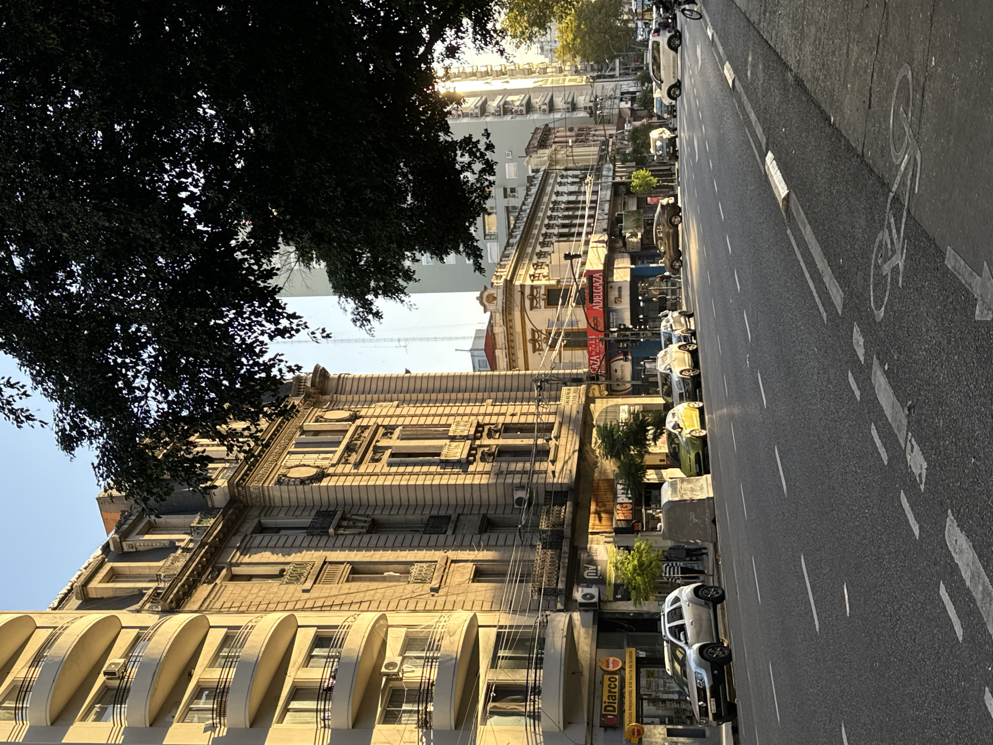 This media file presents a vibrant, sunlit street scene in Buenos Aires, Argentina, captured during what appears to be the golden hour, either early morning or late afternoon, evidenced by the warm light and long shadows. The image is taken from a slightly elevated perspective, looking down a bustling urban street lined with diverse architectural styles.
On the left, a large, dark-leafed tree dominates the foreground, partially obscuring the upper sections of the buildings. The buildings themselves are a key subject. The closest building on the left features unique, modern-looking curved balcony structures. Further down, an older, ornate building stands out with classical architectural details, including elaborate window frames, decorative balconies, and a textured facade, hinting at the city's rich history. Beyond these, taller, more contemporary buildings with numerous air conditioning units are visible, some illuminated by the low sun.
The street itself is asphalt, marked with dashed white lines separating traffic lanes. A distinct white bicycle symbol painted on the right side of the road indicates a dedicated bike lane. Numerous cars are parked along the curb and others are visible in motion, suggesting an active thoroughfare. Overhead, a complex network of power lines crisscrosses the scene.
Visible text includes a prominent yellow sign for "Diarco" with the slogan "precios mayoristas, a la vuelta de tu casa." (wholesale prices, around the corner from your house). Further down, red banners display "ADELGAZA," "CAZA 71," and "Arcoiris." Partially obscured signs show "BIGPINS" and "INNMA." The overall scene captures the dynamic atmosphere of a city street, blending historical architecture with modern urban elements under a clear, sunny sky.