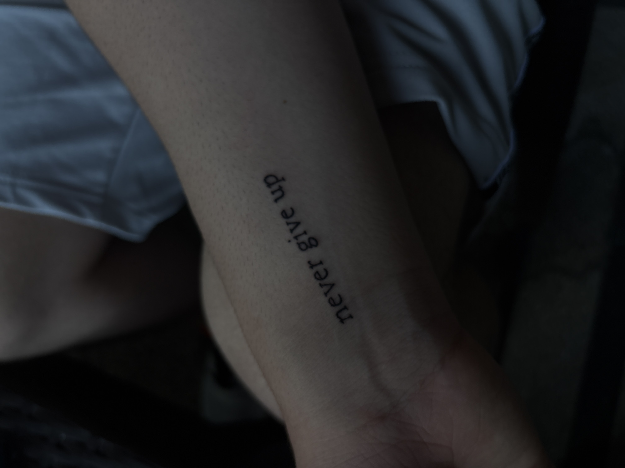 A photograph depicts a person's forearm with a tattoo that reads "never give up" in a simple, sans-serif font. The arm is positioned in the foreground, partially obscuring the subject's leg and a portion of their clothing, which appears to be a light-colored garment. The background is dark and indistinct. There is no visual information to suggest the location is Gotse Delchev, Bulgaria, or to provide any specific details about actions, interactions, or the environment.