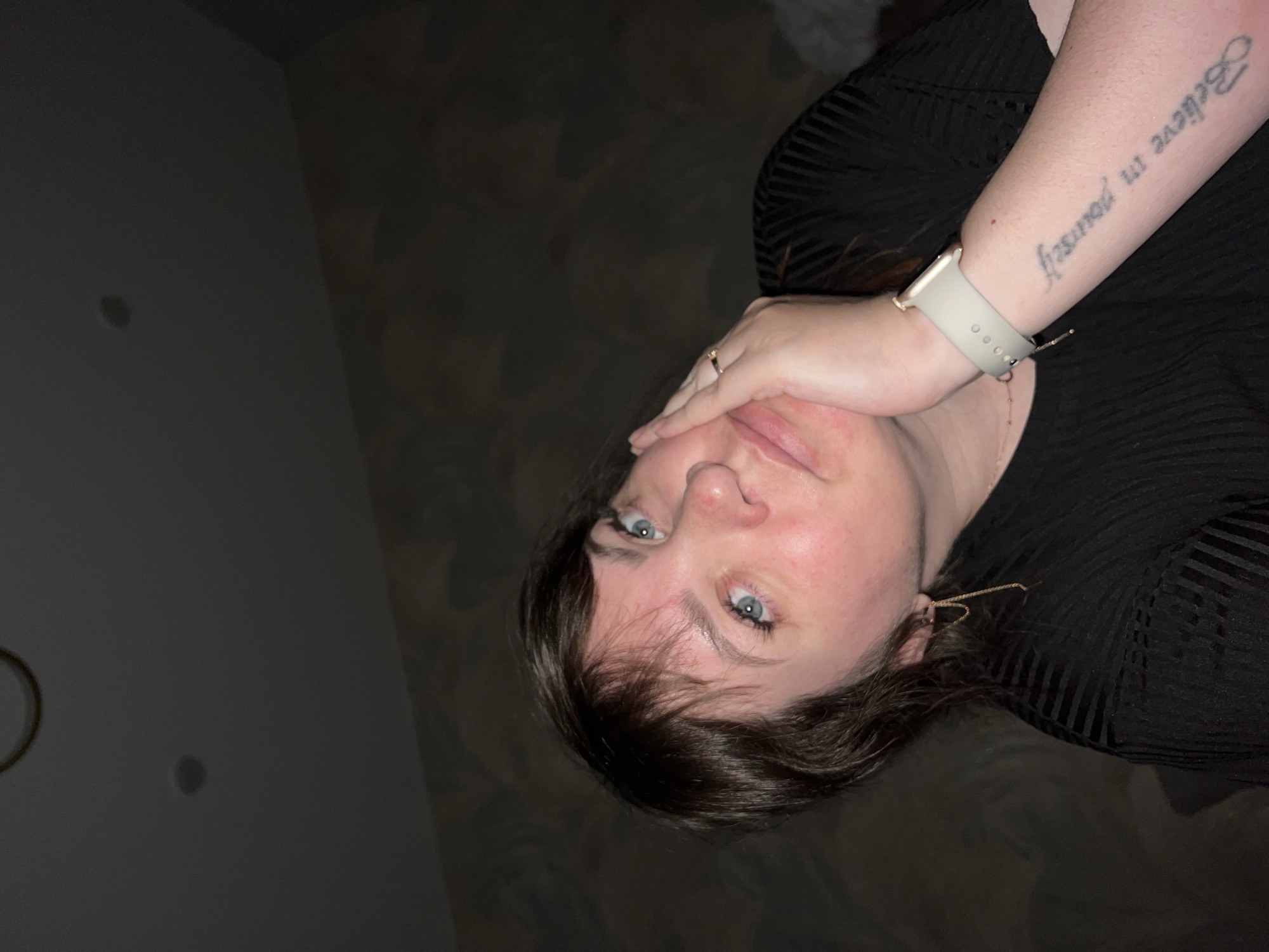 A photograph depicts a woman with blue eyes and dark brown hair. She is wearing a black, horizontally striped shirt. Her left hand, adorned with a ring, is near her face. Her left forearm features a tattoo with the phrase "Believe in yourself" and a smartwatch with a light-colored band. The background is darkly lit and indistinct, with a glimpse of a wall with fixtures. The photograph does not contain information about the city of Noviy, Russia.