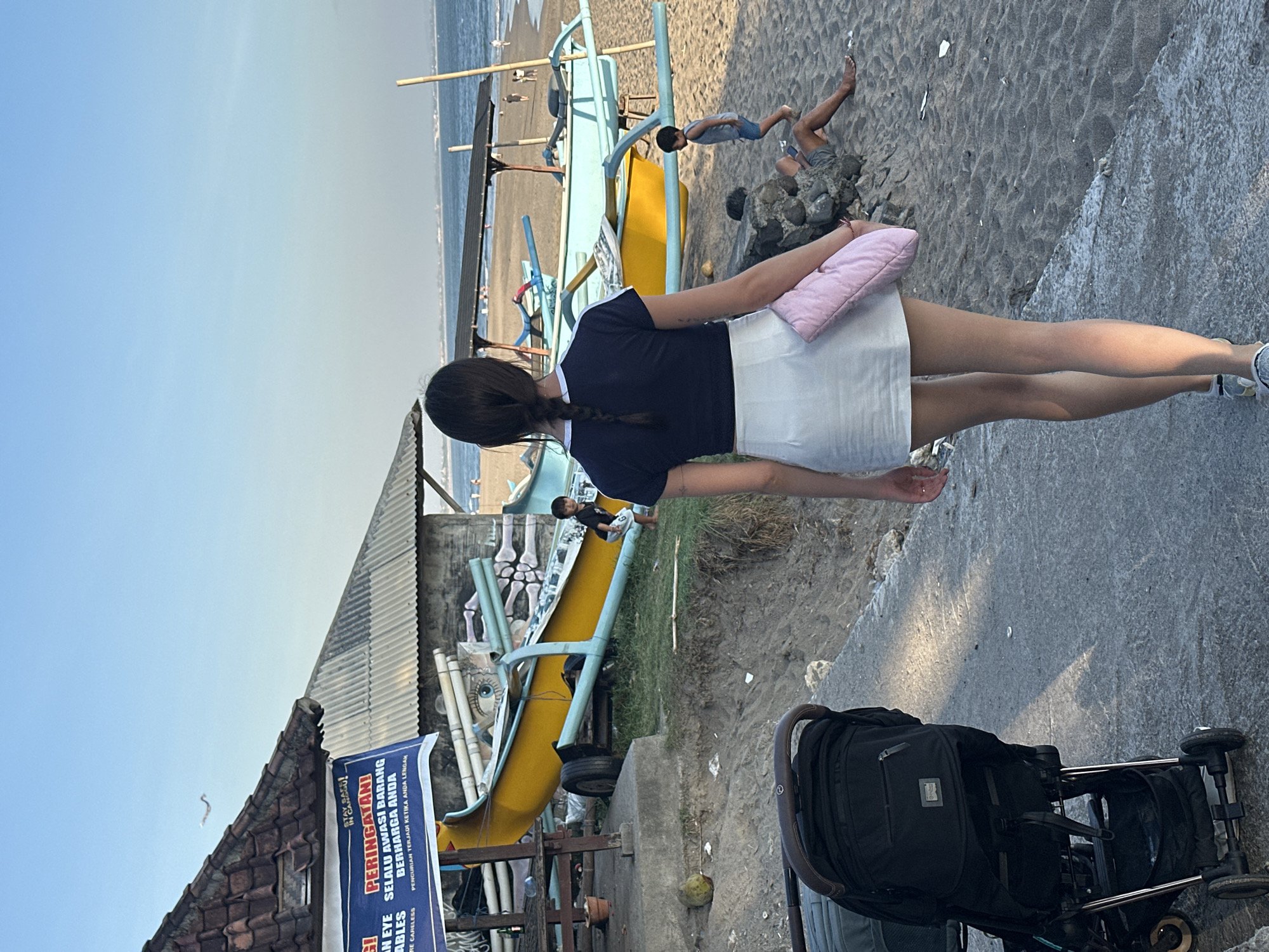 This is a portrait-orientation, full-body shot of a young woman standing on a sandy beach, facing away from the camera. She is wearing a dark blue t-shirt and a white skirt. She is holding a small, light pink pillow in her right arm. Behind her, a yellow and light blue fishing boat is pulled up onto the sand. Several people are visible in the background, some near the boat and others walking on the beach. The ocean is visible in the distance. The sky is clear and blue, suggesting a sunny day, possibly late afternoon due to the long shadows. A baby stroller is parked in the foreground to the right. A sign with Indonesian text is partially visible on the left, reading "PERINGATAN!" which means "WARNING!" and includes phrases about watching belongings. The scene appears to be a casual day at the beach in a populated area, likely a tourist destination given the presence of fishing boats and commercial signage. The overall mood is relaxed and sunny.
