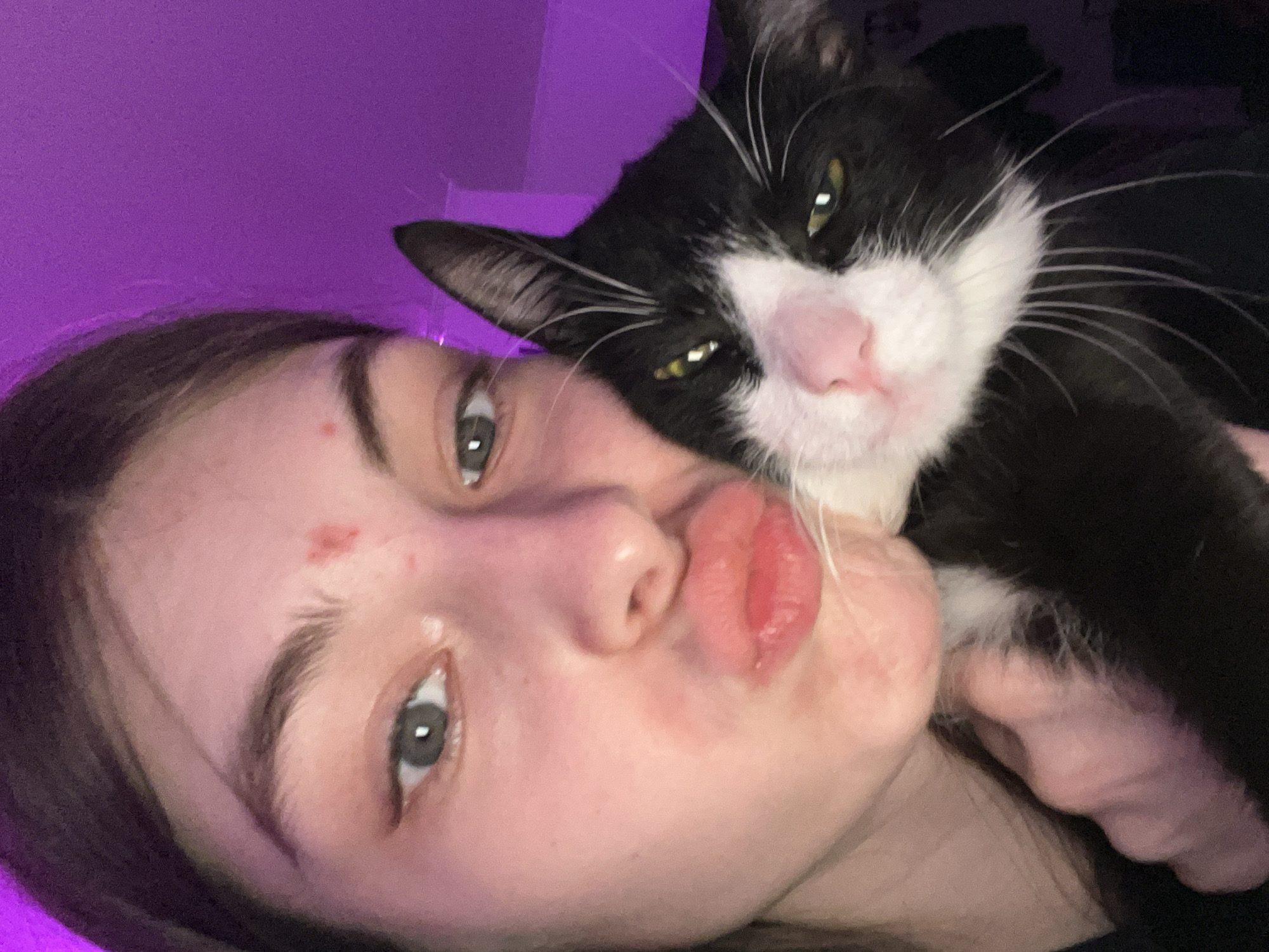 A close-up selfie features a young person and a black and white cat, possibly taken in Saint Petersburg, Russia, as indicated by the provided context. The person is on the left, facing towards the right with their head tilted. They have dark hair, light-colored eyes, and are making a "duck face" or puckering their lips. A small red mark or blemish is visible on their forehead.
The cat, a tuxedo cat with black fur, white markings on its face and chest, and green-yellow eyes, is positioned on the right side of the frame, resting against the person's head or shoulder. Its expression appears somewhat sleepy or indifferent, with its eyes partially closed. The person's left hand is visible, seemingly holding the cat close.
The background is a solid purple color on the left side, suggesting artificial lighting like LED strips, and fades into darkness on the right. The scene is indoors, and the artificial light makes the time of day undeterminable, though it likely appears to be evening or night. There is no visible text or specific location cues within the image itself to confirm the provided location context. The overall mood is affectionate and playful.