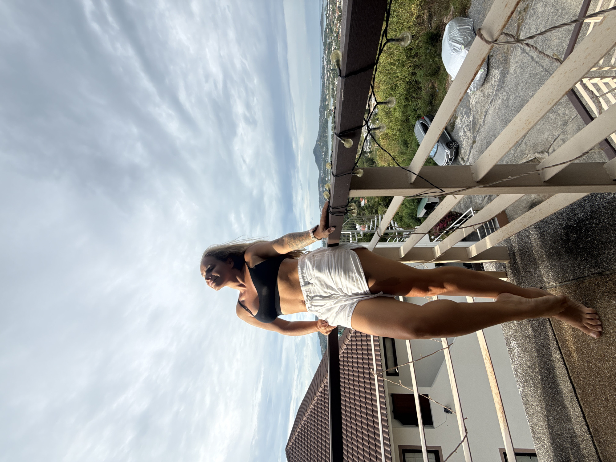 A woman stands on a balcony in the city of Ko Samui District, Thailand. She is wearing a black sports bra and white shorts. Her head is tilted back, eyes closed, and she appears to be basking in the sun. Her right hand rests on the balcony railing, and her left arm is bent at the elbow, with her hand on her hip. The balcony overlooks a residential area with houses and a view of the ocean and hills in the distance. The sky is cloudy. There are string lights hanging along the railing. Two cars are parked below the balcony.