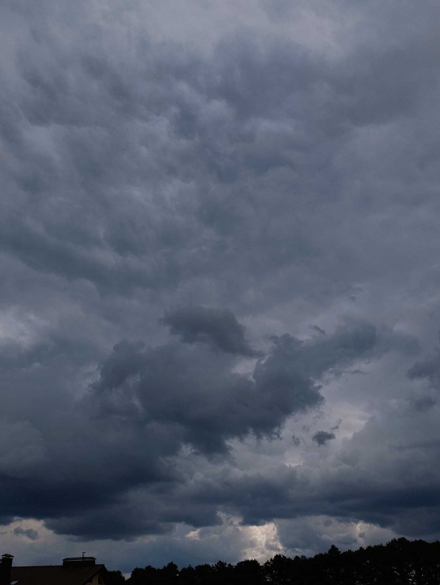 A stormy, overcast sky dominates the image, with heavy, dark gray clouds filling the frame. The clouds exhibit various textures and densities, with some areas appearing lighter and more diffused, suggesting breaks in the cloud cover or distant sunlight. In the lower portion of the image, a dark silhouette of trees is visible, bordering the horizon. A small section of a building's roof, appearing to be made of dark tiles, is visible on the far left, with a chimney. This scene appears to be an outdoor view, possibly from a residential area, under a dramatic and foreboding sky. The location is the city of Minsk, Belarus.