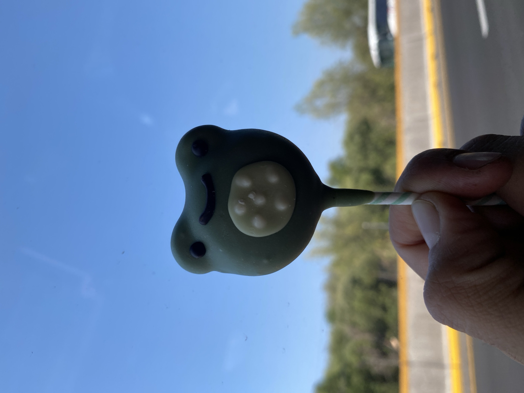 A hand holds a frog-shaped lollipop in front of a window. The lollipop is green with a white belly and has a smiling face. Outside the window, there is a blue sky and trees. A road with a yellow stripe can be seen on the right edge of the image. The location appears to be Santa Ana Xalmimilulco, Mexico.
