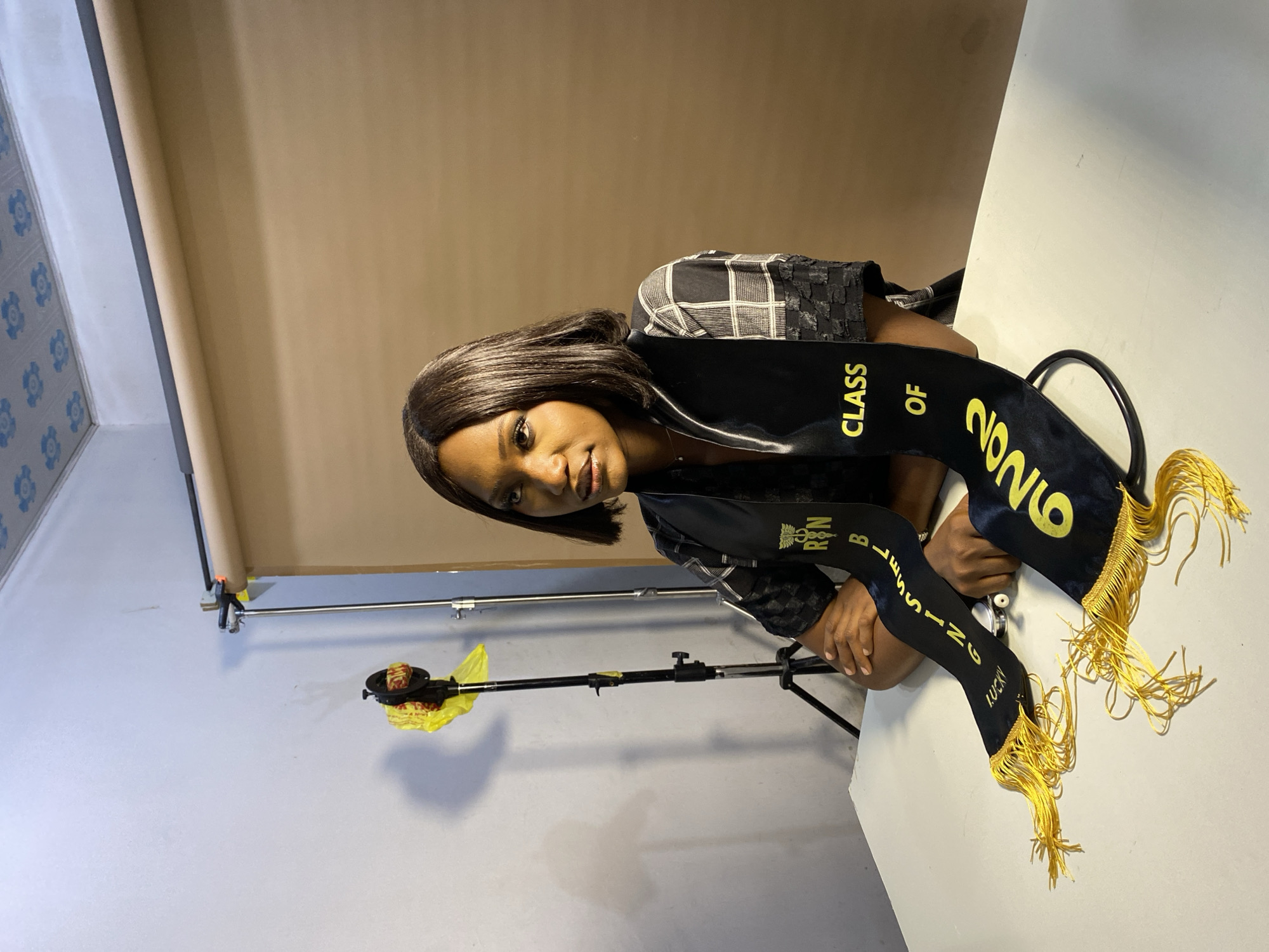 A young woman is posing indoors in a studio setting. She is wearing a black graduation stole with yellow tassels and the text "Class of 2026" and "RN B. Blessing Lucky" printed on it. The background consists of a beige wall and a white surface, likely part of the studio setup. There is photography equipment visible, including a stand with weights and a backdrop support system. Based on the request, the location is specified as Gura Top, Nigeria, although there is no visual information in the image to confirm this.