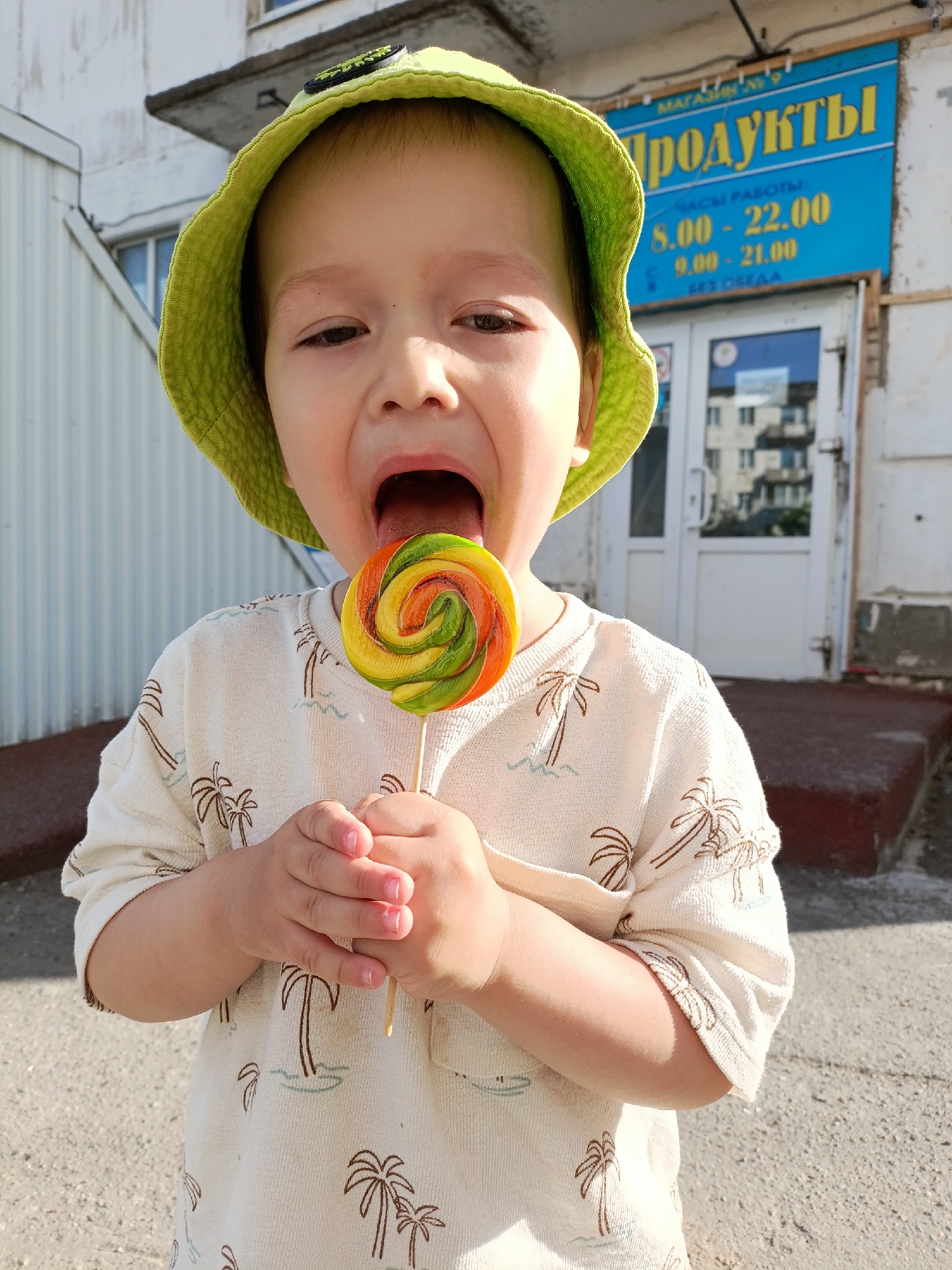 A young child, likely a toddler, is seen outdoors in the city of Chaikovsky, Russia. The child is wearing a light green bucket hat and a cream-colored shirt patterned with brown palm trees. He holds a large, colorful swirl lollipop, which he is in the process of licking, tongue extended. Behind the child is a building with a sign in Russian that translates to "Products". The sign also displays hours of operation. The building's façade appears somewhat worn. The scene is illuminated by natural daylight.