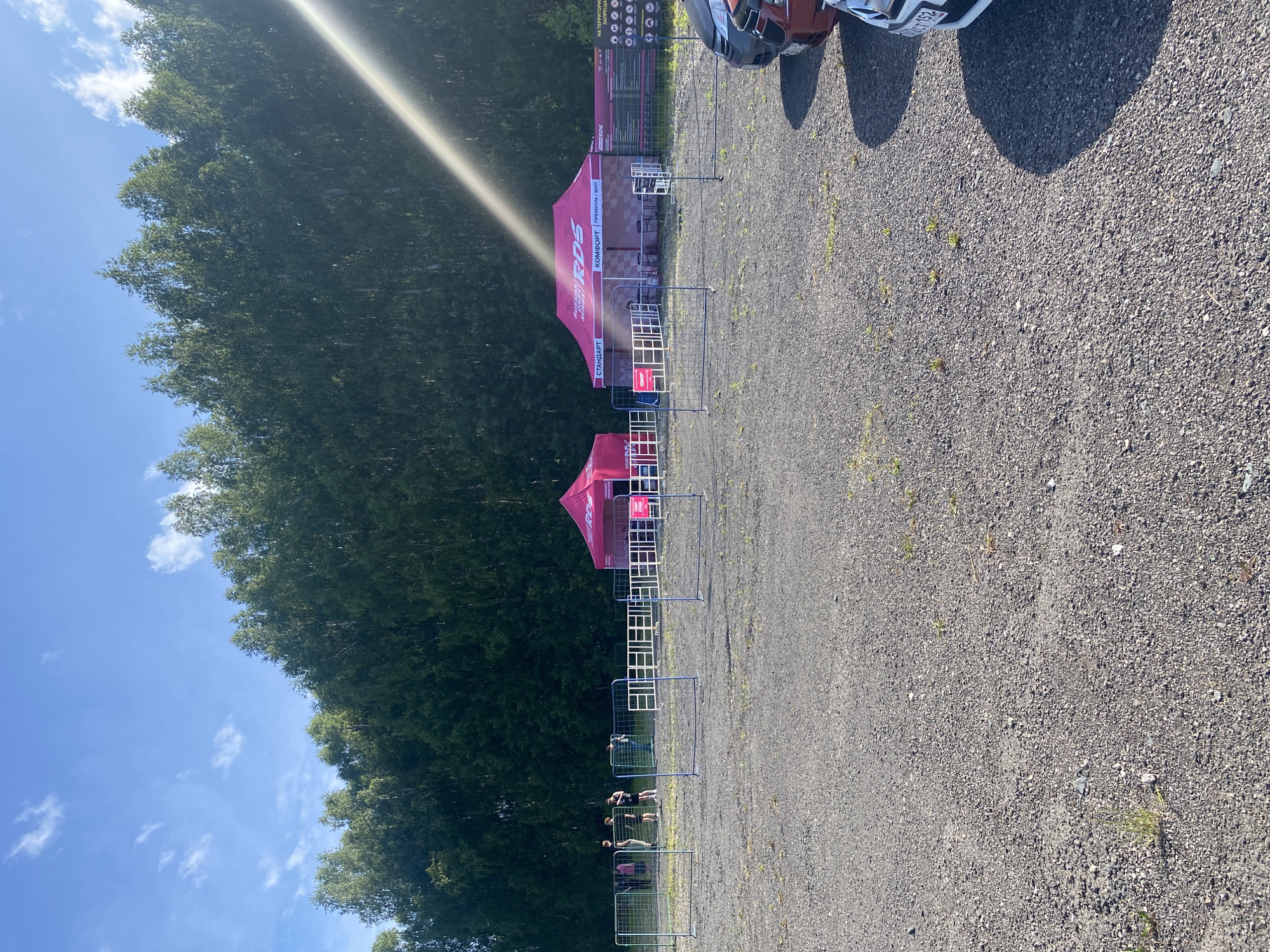 In Berëzovka, Russia, on a clear day with a bright blue sky and scattered clouds, a large group of trees forms a dense, green backdrop. In the foreground, a gravel-covered lot is divided by temporary metal fencing. Two pink tents with white lettering, appearing to be vendor or information booths, are set up behind the fencing. The tents have "RDS" and "RUSSIAN" visible on them. A few people are gathered near the tents and within the fenced area. Cars are parked on the right side of the image, partially visible. The overall scene suggests an outdoor event or gathering.