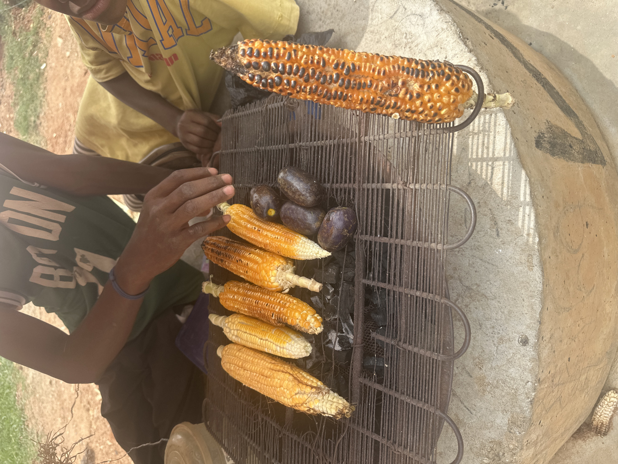 In Gwarinpa, Nigeria, this image captures a roadside food stall where corn and what appear to be small eggplants are being grilled over charcoal. Two young people, likely a boy and a girl, are present. The boy, wearing a green t-shirt with "BOM" visible, is positioned on the left, with his dark hands reaching towards the grill. A blue rubber band is on his left wrist. The girl, on the right, is wearing a yellow t-shirt with orange lettering and is looking towards the food. Several ears of corn are arranged on a metal grill rack, some showing char marks from the heat. Beside the corn are several dark, oval-shaped items, possibly eggplants. One ear of corn is placed vertically next to the grill, held by a metal stand. The stall is set up on a concrete surface, with dry grass and dirt visible in the background, suggesting an outdoor, possibly dusty environment.