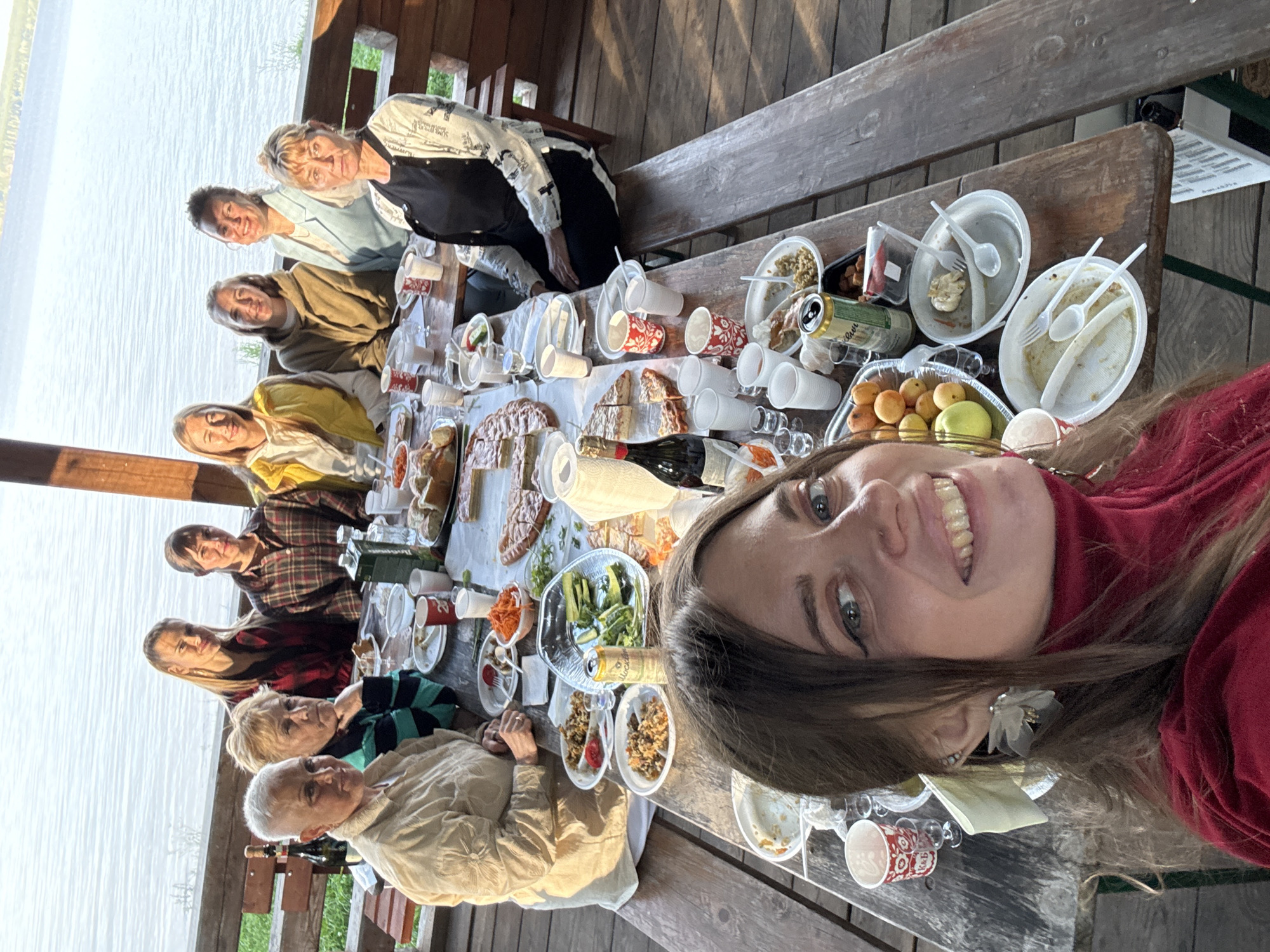 A group of nine women is gathered around a wooden table laden with food and drinks, seemingly for a meal outdoors by a body of water. The table is set with plates, cups, bottles, and various dishes, including salads, pies, and what appears to be rice. Some women are looking at the camera, while others are engaged in conversation or eating. The setting suggests a casual, social gathering in a rustic environment, possibly a park or a picnic area, with a lake or river visible in the background.