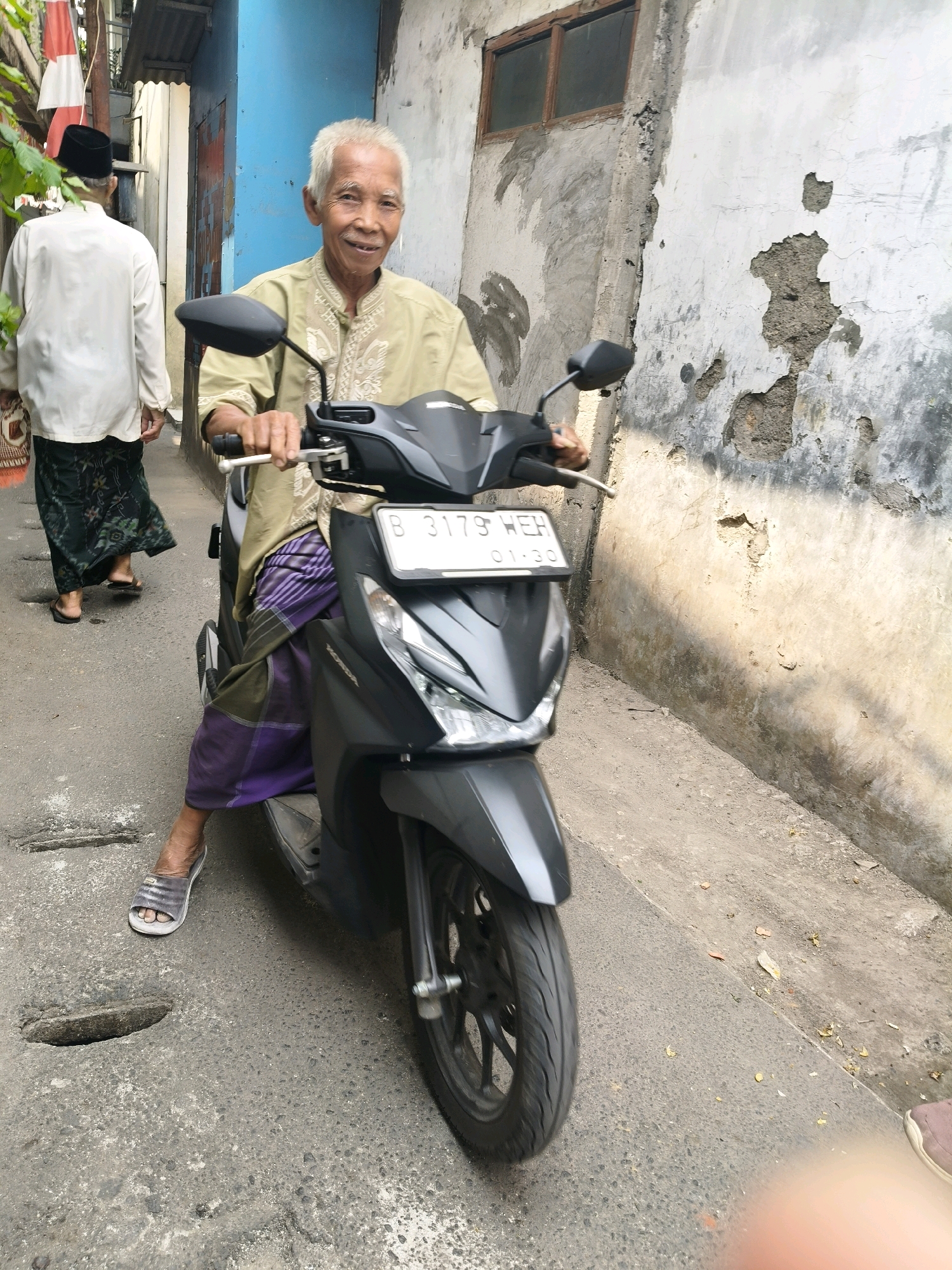 An elderly Indonesian man with grey hair and a light green shirt adorned with white embroidery is seated on a dark grey motorcycle. He is wearing a purple and green sarong and grey sandals. The man is looking towards the camera and smiling. Behind him, another man wearing a white shirt and a black cap walks away from the camera, also wearing a patterned sarong and sandals. They are in a narrow alleyway with a rough concrete wall on the right, displaying peeling paint and exposed concrete. A blue building is visible to the left, with a corrugated metal roof and a red and white flag hanging. The ground is paved with worn asphalt, featuring visible cracks and potholes. The license plate on the motorcycle reads "B 3179 WEH". This scene is typical of a residential area in South Tangerang, Indonesia, characterized by its modest buildings and narrow, often uneven, street infrastructure.