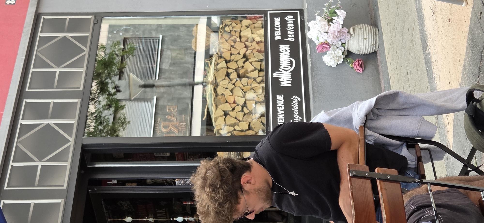 A man with curly brown hair and sunglasses is sitting at a table outside a business. He is wearing a black t-shirt and a silver necklace. The business has a large window with a sign that reads "Bienvenue", "willkommen", "WELCOME", and "benvenuto". Inside the window, there is a display of firewood. To the right of the sign, there is a vase with pink and white flowers. The man is looking down and to his left. The environment appears to be an outdoor seating area of a business. The image is taken during the daytime.