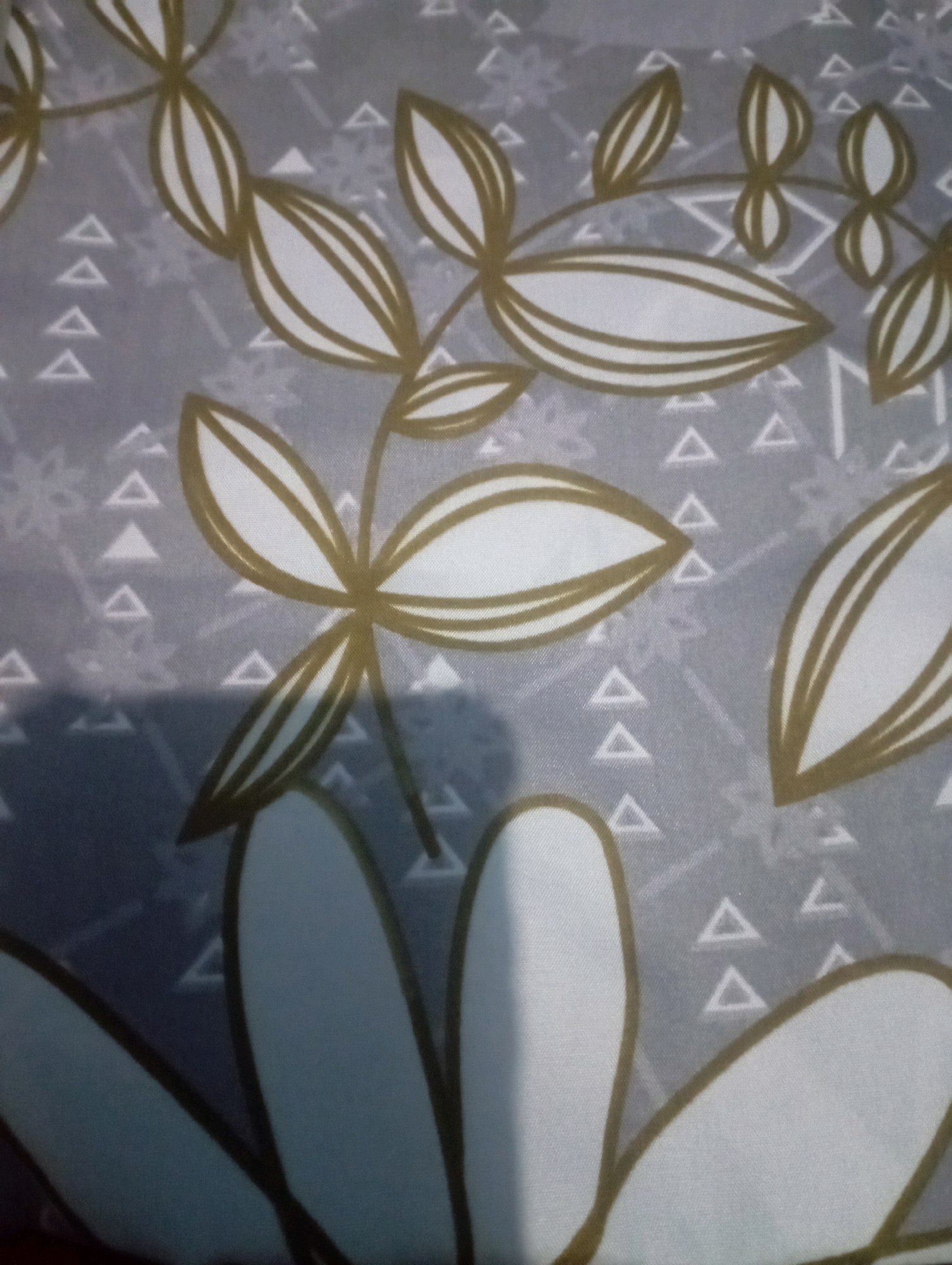The image displays a close-up view of a fabric with a repeating pattern. The pattern features stylized golden-brown leaves or petals outlined in dark brown, set against a light grey background with a subtle geometric design of small white triangles and faint grey lines. The lighting creates some shadows and highlights on the fabric, suggesting it is a soft material. There are no people, recognizable landmarks, or environmental features visible that would indicate a specific location in Senangsari, Indonesia.