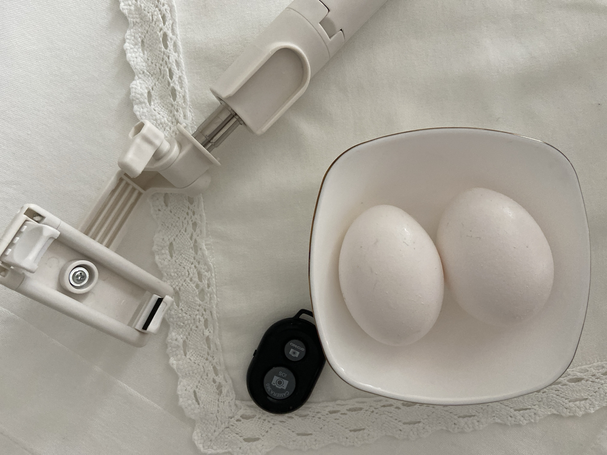 On a white tablecloth with lace edges, a white selfie stick lies next to a small, white, square bowl containing two white eggs. A small, black, oval-shaped remote control rests between the bowl and the selfie stick. The location is not discernible from the provided image.