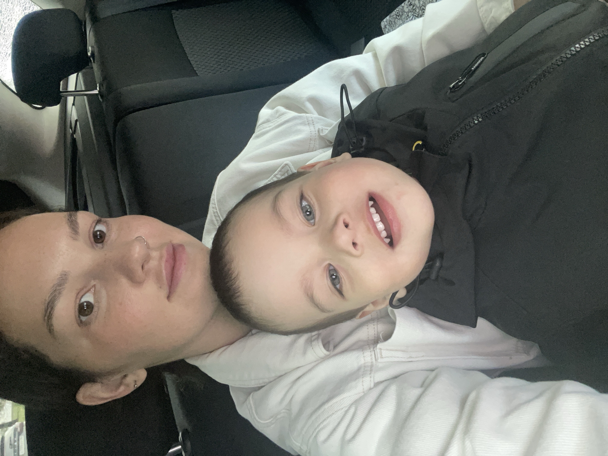 A mother and child are in the back seat of a car, likely in Izhevsk, Russia, judging by the context. The mother, positioned on the left side of the frame, is wearing a light-colored jacket over a white shirt. She has her hair pulled back, and is wearing a nose ring and an earring. She is looking towards the camera, and her left cheek is slightly turned towards the viewer. The child is in front of her, also looking towards the camera, with a wide smile and open mouth, revealing teeth. The child is wearing a black jacket. The background shows the dark interior of a car, with car seats and headrests visible. The image appears to be taken from a selfie perspective, with the mother holding the camera.