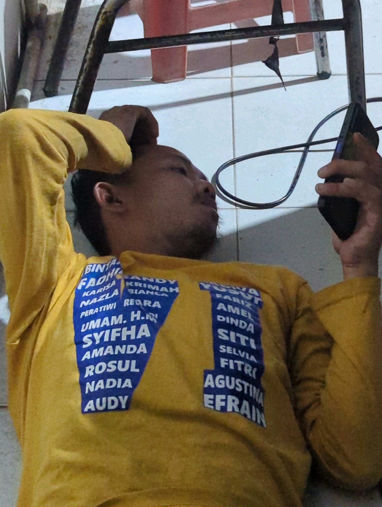 A person wearing a yellow long-sleeved shirt lies on a tiled floor, holding a black smartphone in their right hand. The shirt has a blue rectangular patch on the chest with text in white. The text is a list of names, with some names partially obscured. The person is resting their head on their left arm, which is bent at the elbow and resting on the floor. A dark colored cord is visible on the floor behind the person. The environment appears to be indoors, with a portion of a metal chair leg and a red plastic object visible in the background. The lighting suggests it is daytime. This image was taken in the city of Batu Belaman, Indonesia.