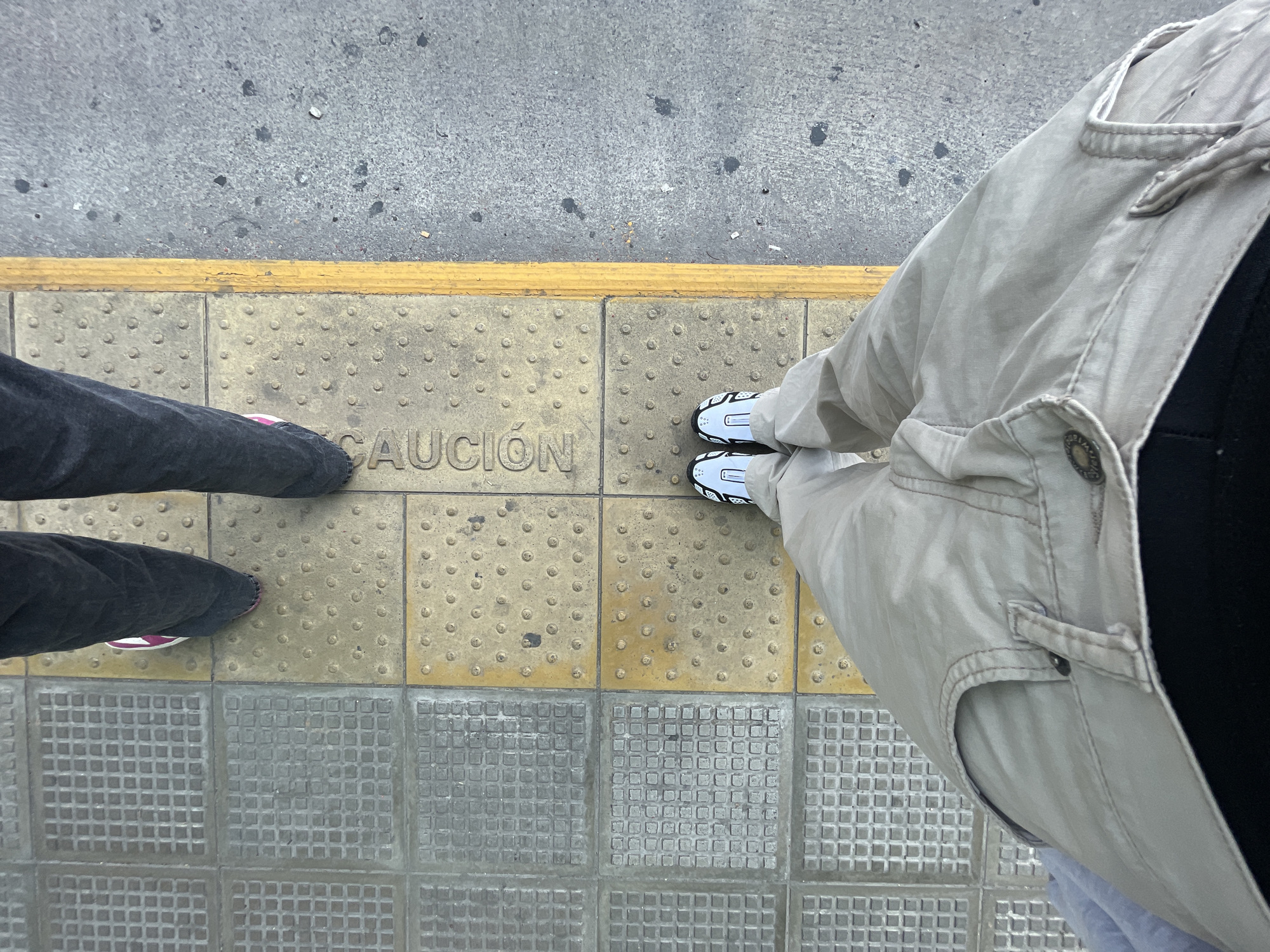 Two people stand on a tactile paving surface marked with "CAUCION" (Spanish for "CAUTION"), bordered by a yellow safety line and adjacent to standard pavement. One individual wears black jeans and sneakers with maroon accents. The other person wears a khaki jacket and white sneakers with black detailing. The scene is likely in Buenos Aires, Argentina, due to the Spanish warning sign. The tactile paving indicates a potentially hazardous area, likely near a transit platform.