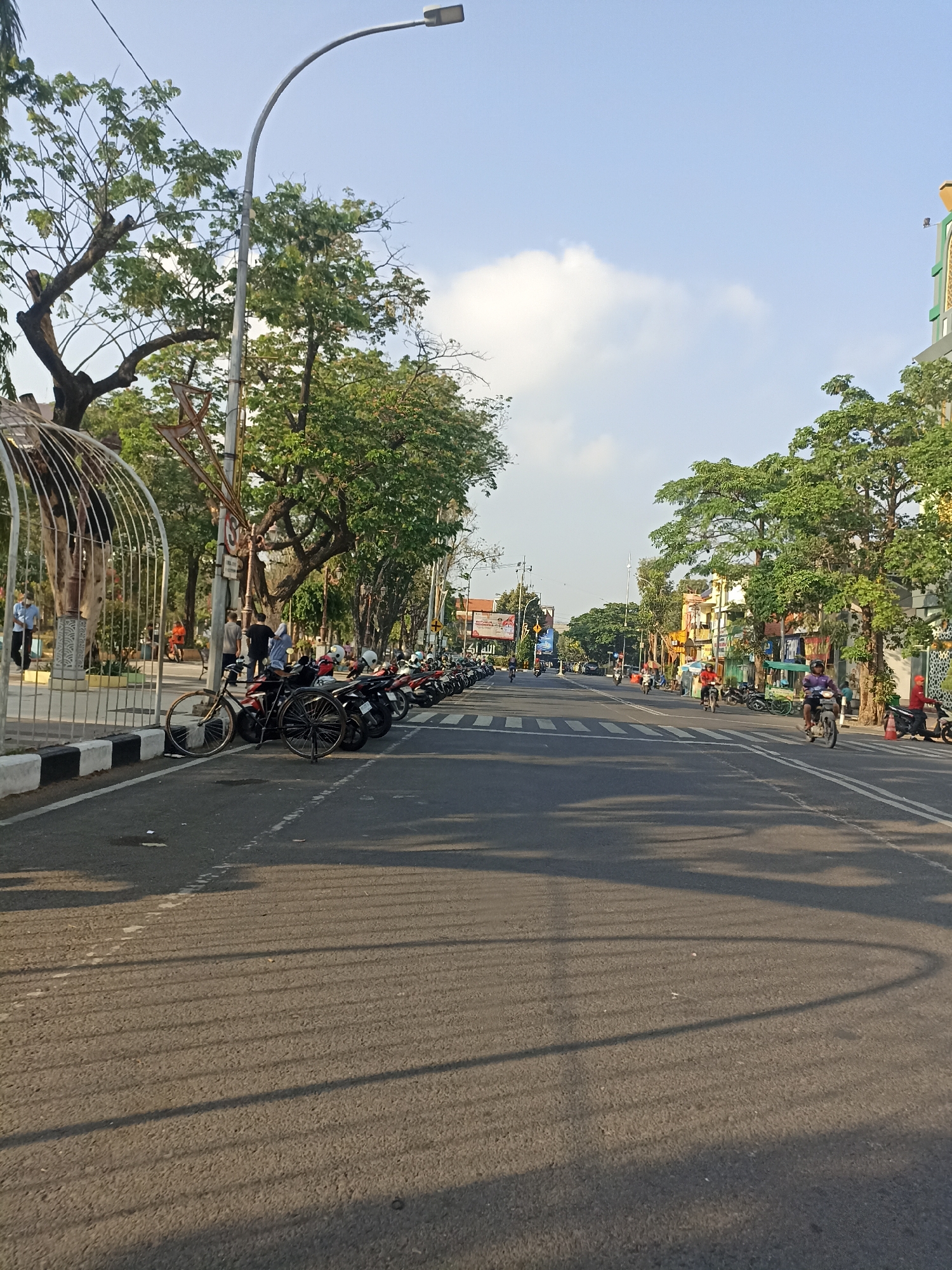 The image depicts a street scene in Lamongan, Indonesia, under a clear sky with scattered clouds. The street is paved and has white lane markings. On the left side of the road, there is a sidewalk with black and white striped curbing. A metal fence, part of a park or public area, encloses trees and some architectural elements. A row of parked motorcycles lines the sidewalk. Two bicycles are parked in front of the motorcycles. Several people are visible on the sidewalk and in the distance, walking or standing. The road itself has light traffic, with a few motorcycles and their riders visible. Trees with dense green foliage line the sides of the street, casting shadows on the road surface. Buildings are visible in the background on the right side of the street, appearing to be shops or commercial establishments. A street light pole with a modern design is also prominent on the left side of the image. The overall impression is a bustling urban environment during daylight hours.