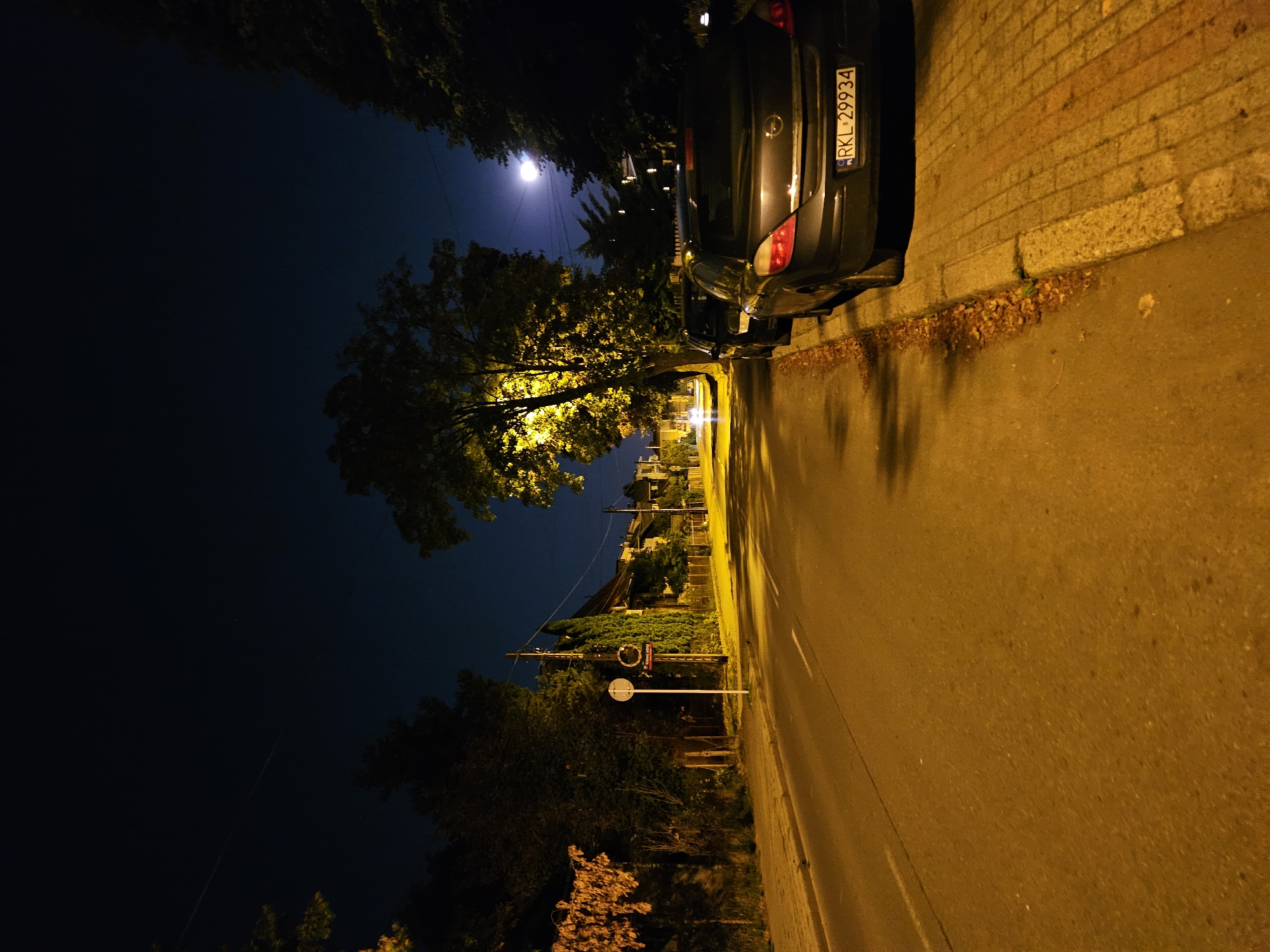 The image depicts a nighttime street scene in Rzeszów, Poland. A dark-colored sedan, identified by its license plate RKL-29934, is parked on the right side of the asphalt road, adjacent to a paved sidewalk. Fallen leaves are scattered along the curb. Further down the street, the lights of buildings and streetlights illuminate the scene, casting a warm glow on the road and pavement. Trees line the left side of the road, their leaves illuminated by artificial light. The sky above is dark with a faint glow suggesting a moon or distant light source. A traffic sign is visible on the left side of the road. The overall impression is that of a quiet residential street at night.