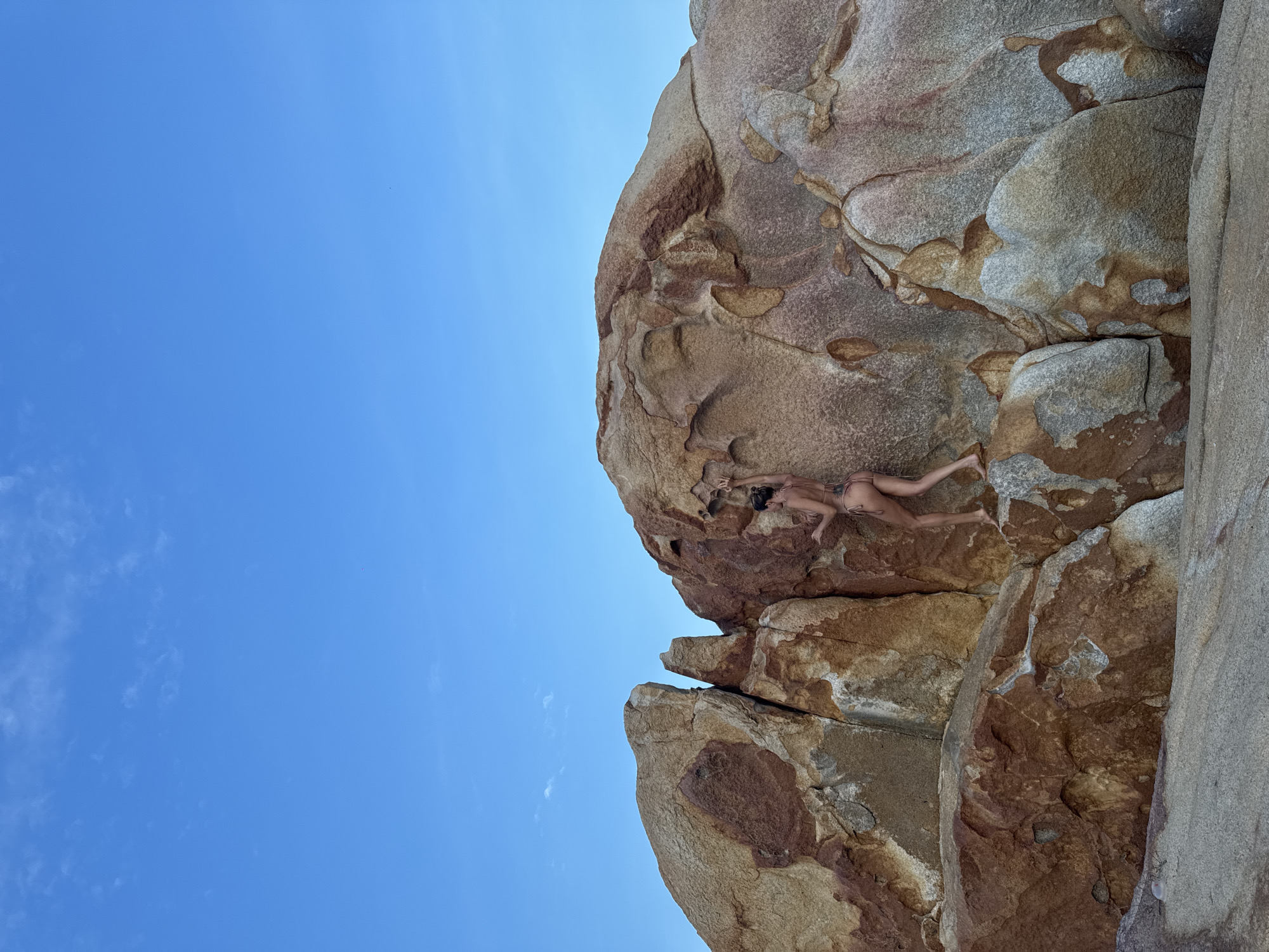 In Thuận Thành, Vietnam, under a clear blue sky with wispy clouds, a woman in a bikini is climbing on a large, multi-colored rock formation. The rocks are predominantly shades of brown, orange, and grey, with visible textures and patterns. The woman is positioned on the right side of a crevice between two large rocks, with her body angled towards the rock face. Her right hand is raised, gripping a protrusion on the rock, and her left hand is also extended upwards. Her legs are bent, with her right foot planted on a ledge and her left leg extended. The landscape appears natural and rugged, with the rocks forming a significant portion of the scene.