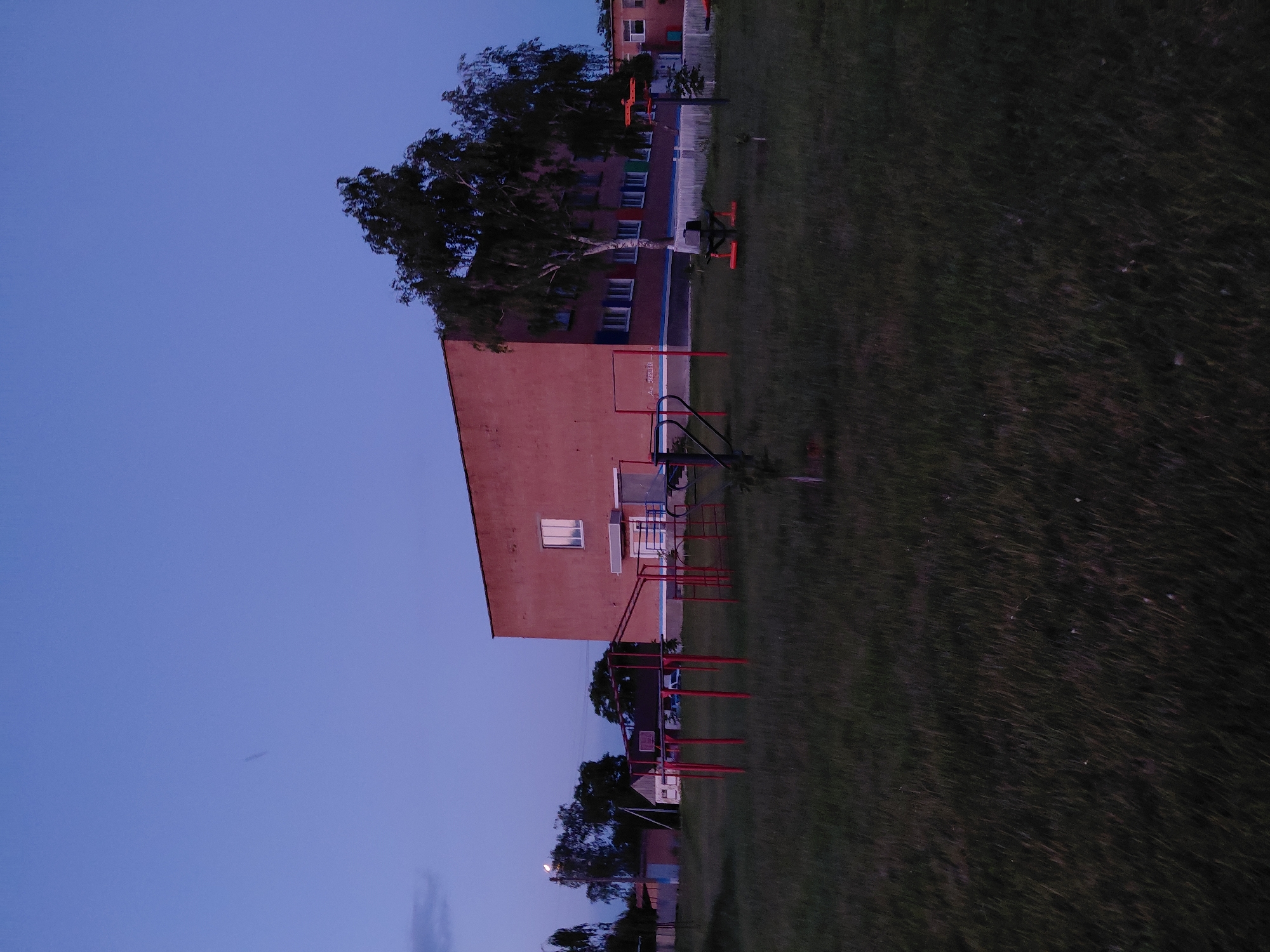 The image depicts an outdoor scene at dusk or twilight, likely in Ustyvytsia, Ukraine. A large, reddish-brown building with numerous windows dominates the background. In front of the building is a grassy area with various pieces of outdoor exercise equipment, including red bars and a black apparatus with orange bases. Trees are visible to the right of the building and in the mid-ground, with some foliage obscuring parts of the structure. To the left of the main building, smaller structures and trees are present. The sky is a deep blue, suggesting a late evening or early morning hour. The overall impression is of a public space, possibly a school or community center, during off-hours. There are no people visible in the image.