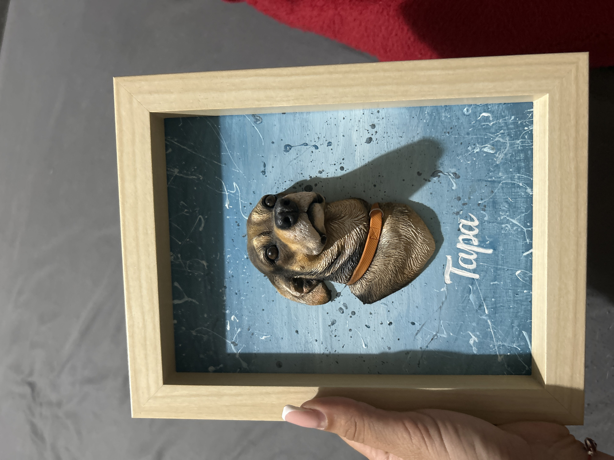 A framed 3D portrait of a dog is held by a person's hand. The portrait features a dog's head and upper chest, with a brown and tan coat and an orange collar. The background of the portrait is a textured blue surface with white splatters and streaks, and the dog's name, "Tapa," is written in white cursive below the dog's chest. The frame is made of light-colored wood. The person's hand is on the left side of the image, with manicured nails. The background appears to be a dark gray surface, possibly a blanket or fabric. A red object is visible on the right side of the image. The lighting casts a shadow from the portrait onto the background.