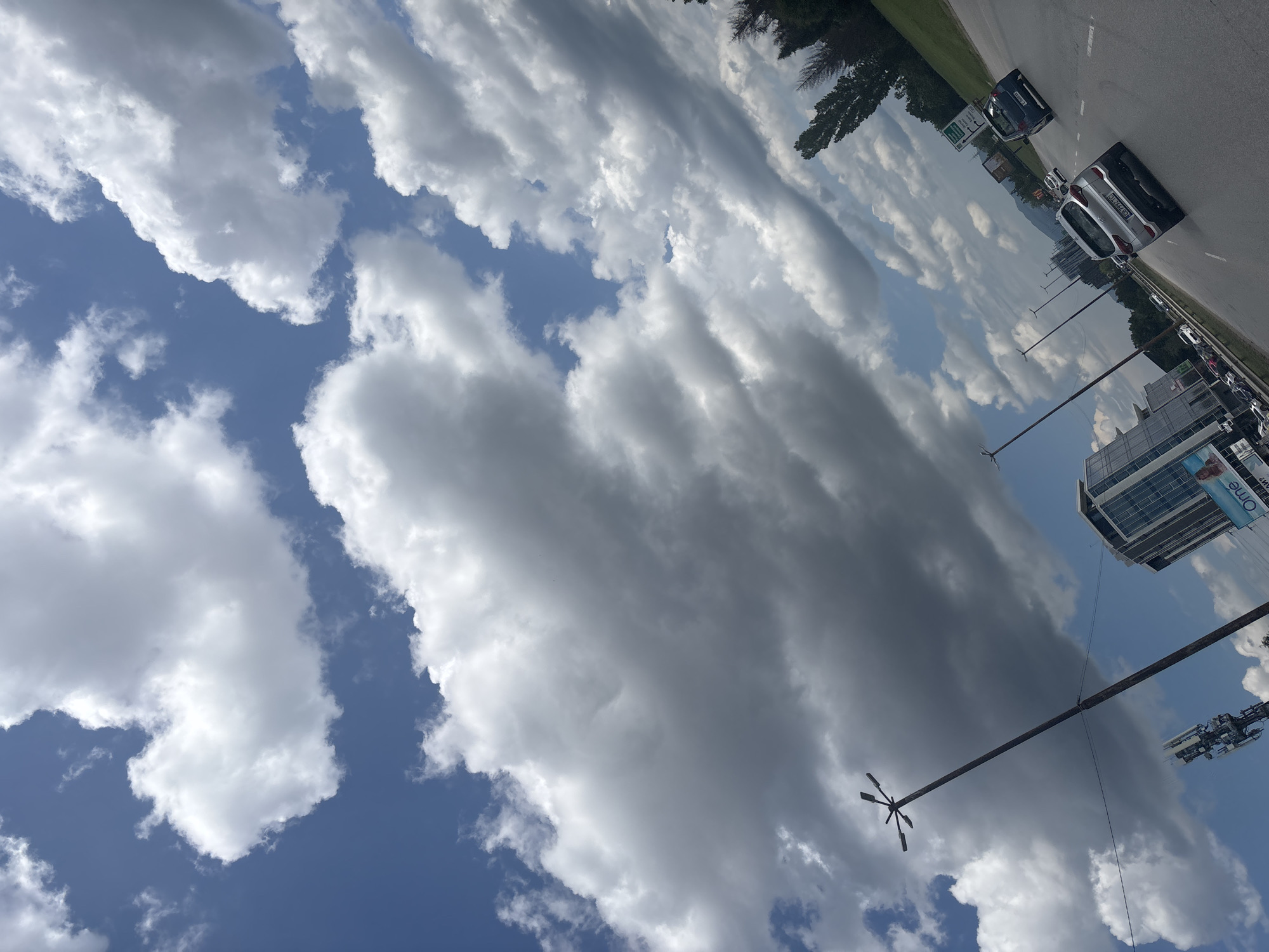 The image captures a slightly tilted, dynamic view of a multi-lane urban thoroughfare under a sky filled with a mixture of bright white and darker grey cumulus clouds. The scene is illuminated by strong daylight, casting soft shadows.
In the foreground, a silver SUV (Hyundai Tucson or similar) is prominently featured, traveling away from the viewer on the right side of the paved road. Its Bulgarian license plate, "CB 5737 KC," clearly indicates Sofia as its registration city. Further down the road, a dark blue hatchback follows, with other vehicles visible in the distance. The road itself has multiple lanes, suggesting a major boulevard.
On the left side of the image, a tall, dark street light pole with a star-shaped array of fixtures extends vertically into the sky. Below and to its left, a modern glass-facade building, possibly an office complex, rises with a large commercial sign or billboard on its lower section that appears to read "Ome" or "Omega." Further left, a cellular tower with antennas is visible against the sky.
To the right of the road, a green embankment is lined with numerous trees, including both tall, slender deciduous trees and some darker coniferous ones, indicating roadside landscaping. In the distant background, faint outlines of hills or mountains are visible under the cloud-dotted sky.
Based on the visible architecture, the multi-lane road infrastructure, and particularly the "CB" Sofia license plate, this scene is located in Sofia, Bulgaria, likely along a major boulevard lined with commercial buildings and office complexes.