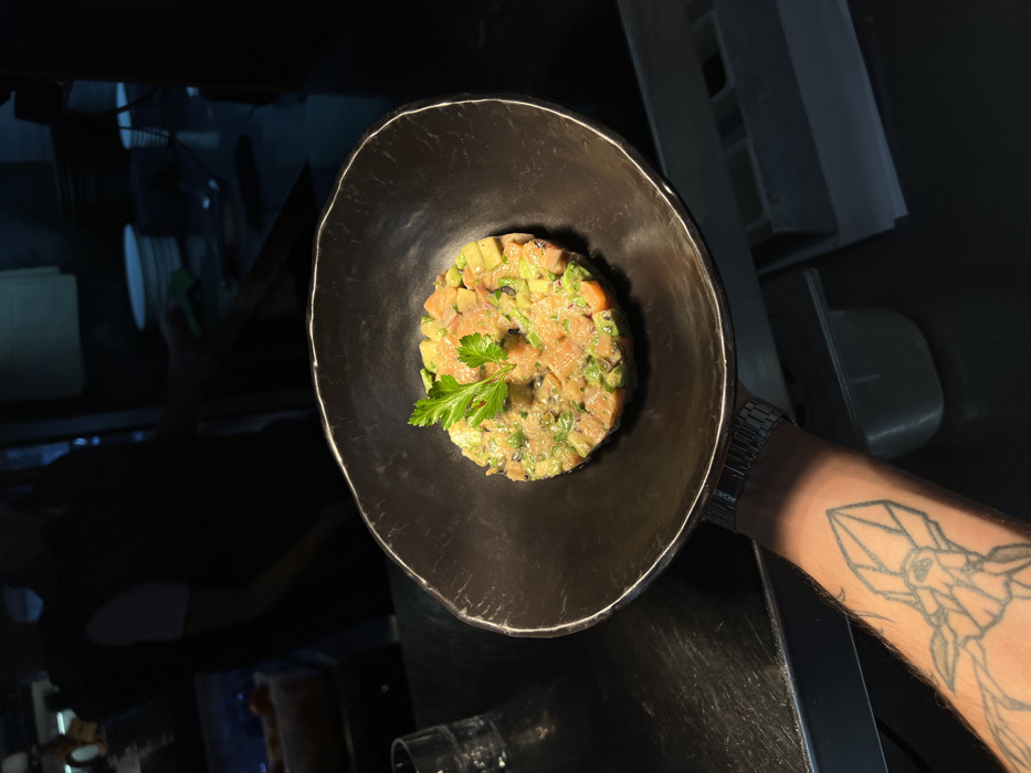 A black bowl contains salmon tartare garnished with a sprig of parsley. The tartare appears to be mixed with diced avocado and other seasonings. A man's arm, adorned with a wristwatch and geometric tattoo, holds the bowl. The background is dark and obscured, suggesting an indoor or low-light setting, possibly a restaurant. The composition is from an overhead perspective, highlighting the food's presentation.