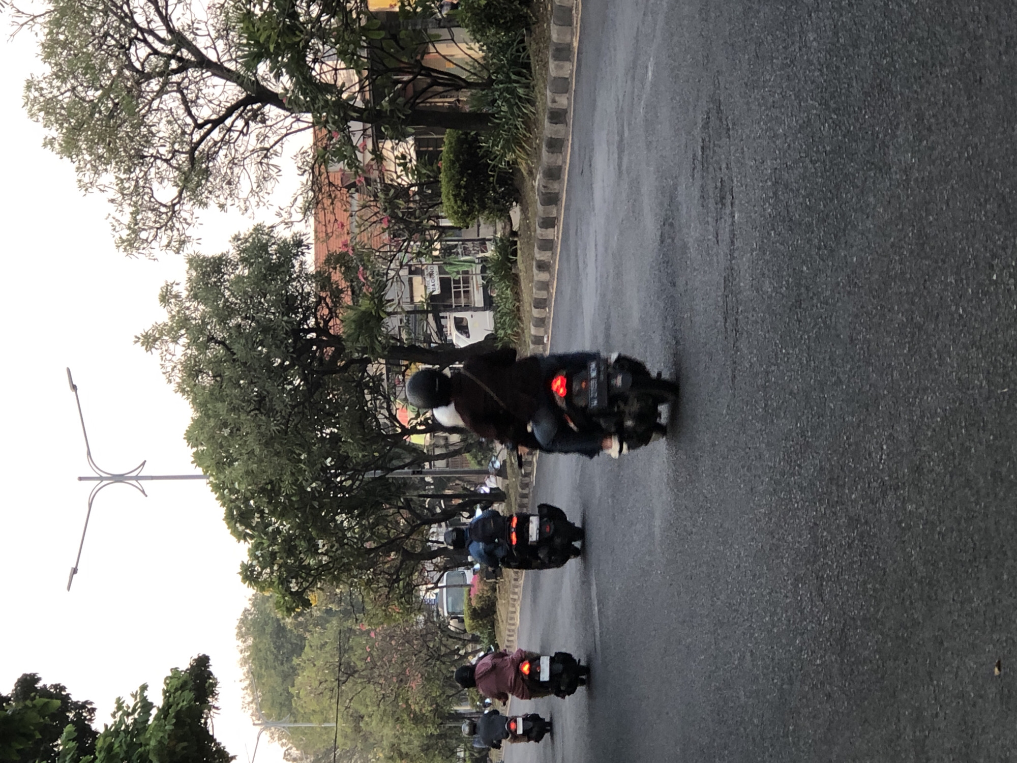 A group of people are riding motorcycles down a paved road in Sumorame, Indonesia. The road has a curb and sidewalk with trees and buildings lining the side. The sky is overcast and the lighting suggests it is either early morning or late afternoon. The people on the motorcycles are wearing helmets and casual clothing. One motorcycle in the foreground has a visible license plate. The trees have green leaves and some pink flowers. There is a white truck parked on the side of the road.