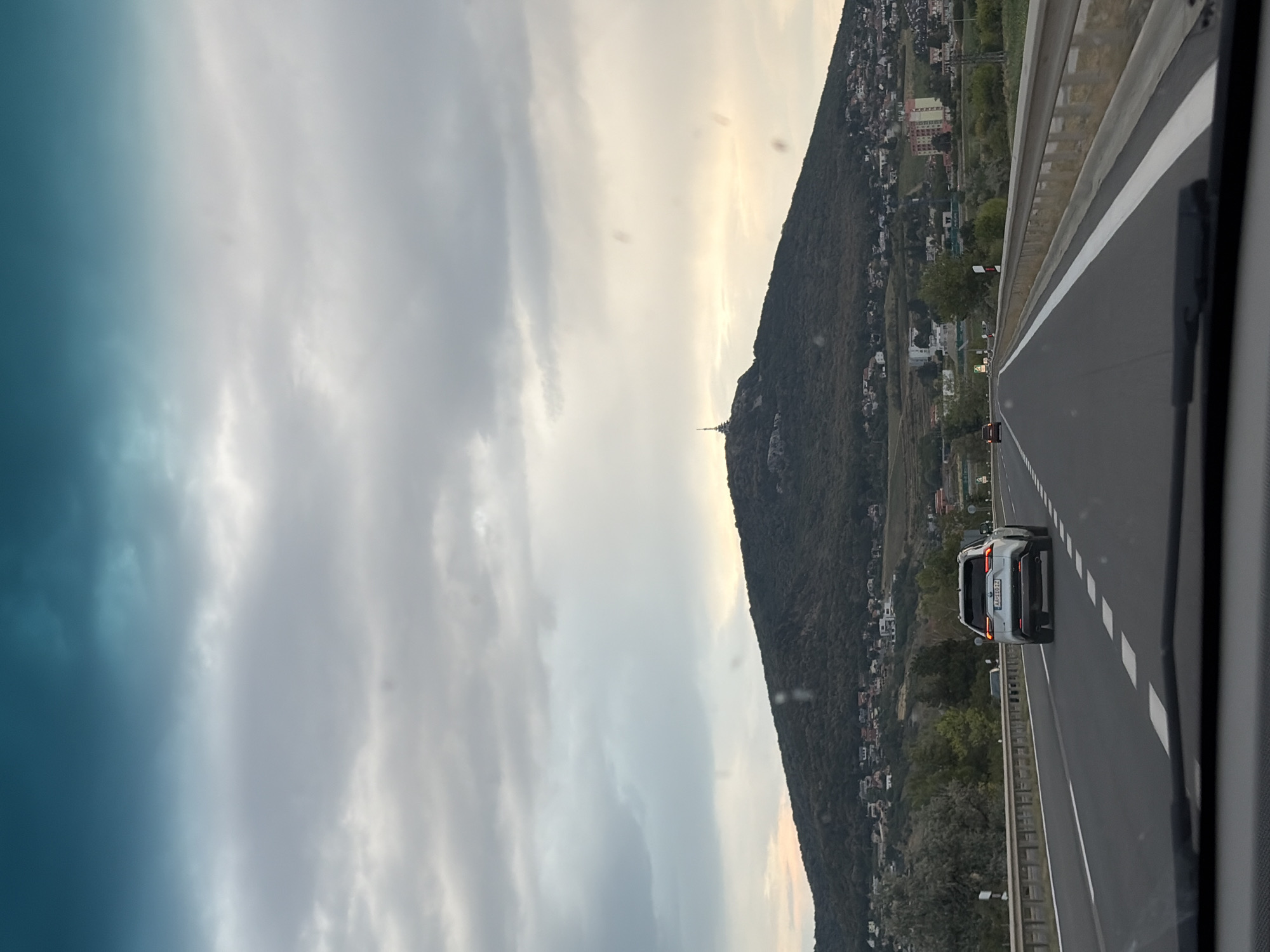 The image shows a view from a vehicle driving on a highway towards a large, forested hill. The sky is overcast with a gradient of teal and grey clouds. On the crest of the hill, a tall radio tower is visible. The slopes of the hill are dotted with houses and trees, forming a suburban or semi-rural landscape. The highway is multi-lane with white and dashed white lines marking the lanes. A silver SUV is in the right lane ahead of the camera, and other vehicles are visible further down the road. The foreground shows the dashboard and windshield of the vehicle from which the photo is taken. Based on the presence of the radio tower on Zobor hill, this scene is likely a view of the city of Nitra, Slovakia, from the D1 highway approaching the city.