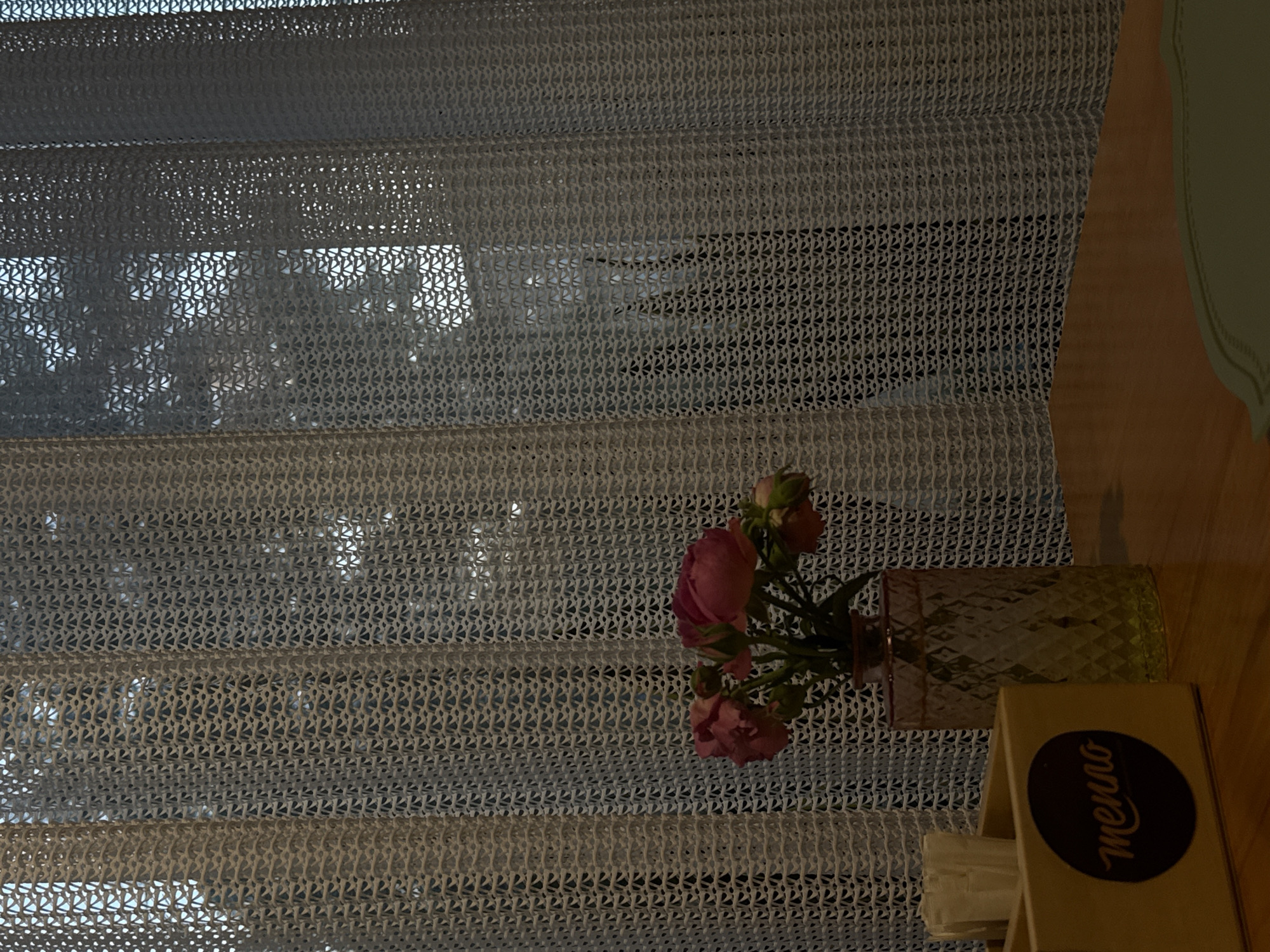 The image depicts a close-up view of a table setting, likely in a cafe or restaurant, near a window covered by a decorative, white, mesh-like curtain. The curtain has a repeating geometric pattern of small, diamond-shaped openings. Through the curtain, the faint silhouettes of trees and possibly buildings are visible, suggesting an outdoor or urban environment.
On the table, a light-colored wooden caddy holds a stack of white napkins. A dark circular logo with the word "menno" in a stylized, script font is affixed to the side of the caddy. To the right of the caddy, a clear glass vase with a diamond-patterned texture holds a small bouquet of pink roses. The vase has a subtle yellow tint. The table surface is a warm, polished wood.
The lighting appears to be dim, possibly indicating an indoor setting in the evening or a shaded area. The overall impression is one of a quiet, possibly intimate, atmosphere. There are no people visible in the image. The location is specified as the city of Abinsk, Russia, which is not evident from the visual information provided within the image itself.