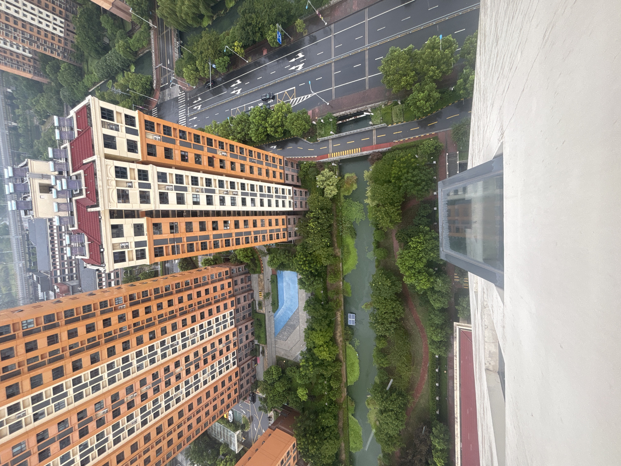An aerial, high-angle shot captures a serene urban landscape in Suzhou Industrial Park, China. The focal point is a modern residential complex characterized by tall, orange-hued buildings with numerous windows. In front of these buildings, a strip of lush green trees lines a calm, dark-colored waterway. A vibrant, rectangular blue swimming pool is nestled amongst the trees, its blue surface contrasting with the surrounding greenery. Beyond the waterway and trees, a multi-lane asphalt road stretches out, wet from recent rain, reflecting the overcast sky. The road is marked with white lane lines and directional arrows, indicating traffic flow. A few dark-colored vehicles are visible on the road. On the right side of the road, more trees create a dense green border. In the background, other buildings and structures, including what appears to be a bridge or overpass, are visible, suggesting a densely populated urban environment. The foreground shows the edge of a modern building, with a geometric glass structure protruding slightly. The overall scene conveys a sense of urban tranquility with a blend of modern architecture and natural elements.