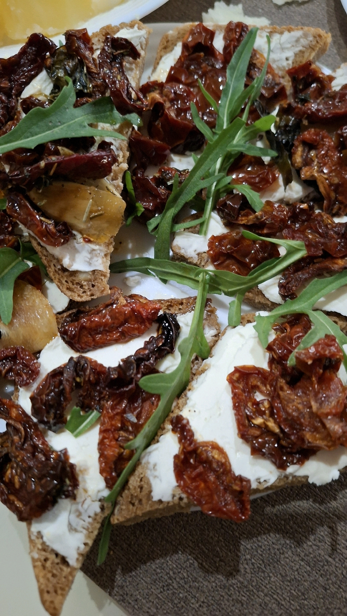 The image displays a close-up, overhead view of a food platter, likely served on a white plate, with a dark brown textured surface visible at the bottom. The platter contains several pieces of what appears to be bruschetta or crostini. Each piece consists of a slice of dark bread topped with a generous spread of white cheese, possibly cream cheese or ricotta. Scattered over the cheese are numerous pieces of sun-dried tomatoes, which are dark red and glossy. Fresh green arugula leaves are also distributed amongst the tomatoes, adding a contrasting color and texture. In the upper left corner, a slice of yellow cheese, perhaps a hard cheese like Parmesan, is partially visible. The food is artfully arranged, suggesting it is a prepared appetizer or snack. The lighting appears to be artificial, casting slight shadows and highlighting the textures of the ingredients. There is no indication of the location within the city of Stowbtsy, Belarus, as the image is solely focused on the food.